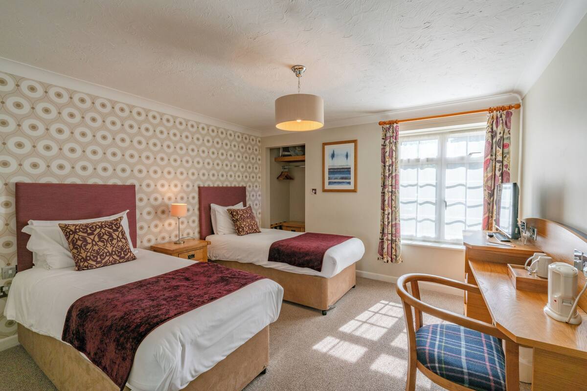 Habitación de hotel con dos camas, un escritorio y una silla. Cuenta con una ventana con cortinas, un cuadro en la pared y una lámpara en el techo.