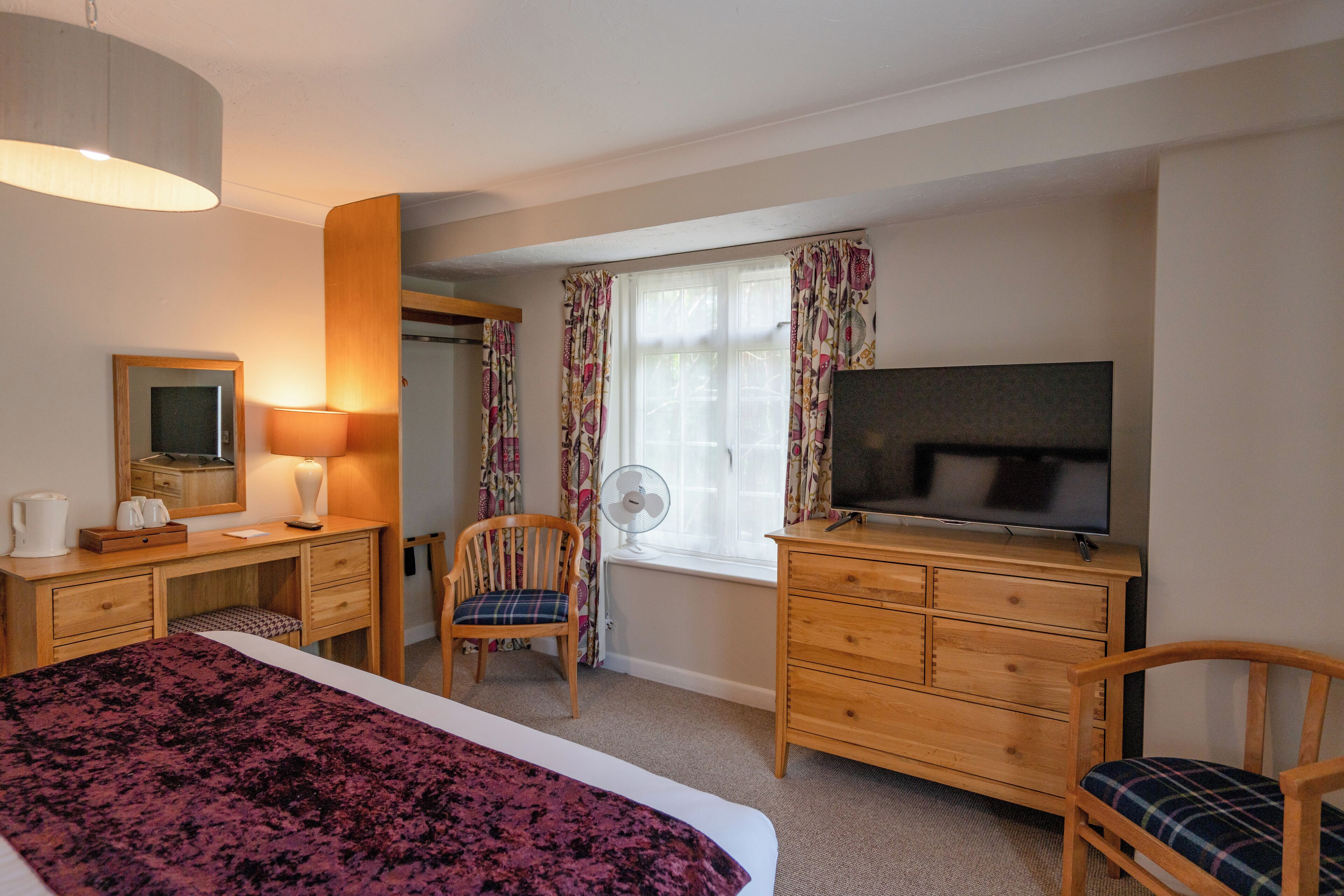 A hotel room with a bed, a desk with a lamp, a chair, a dresser, a TV, and a window with curtains.