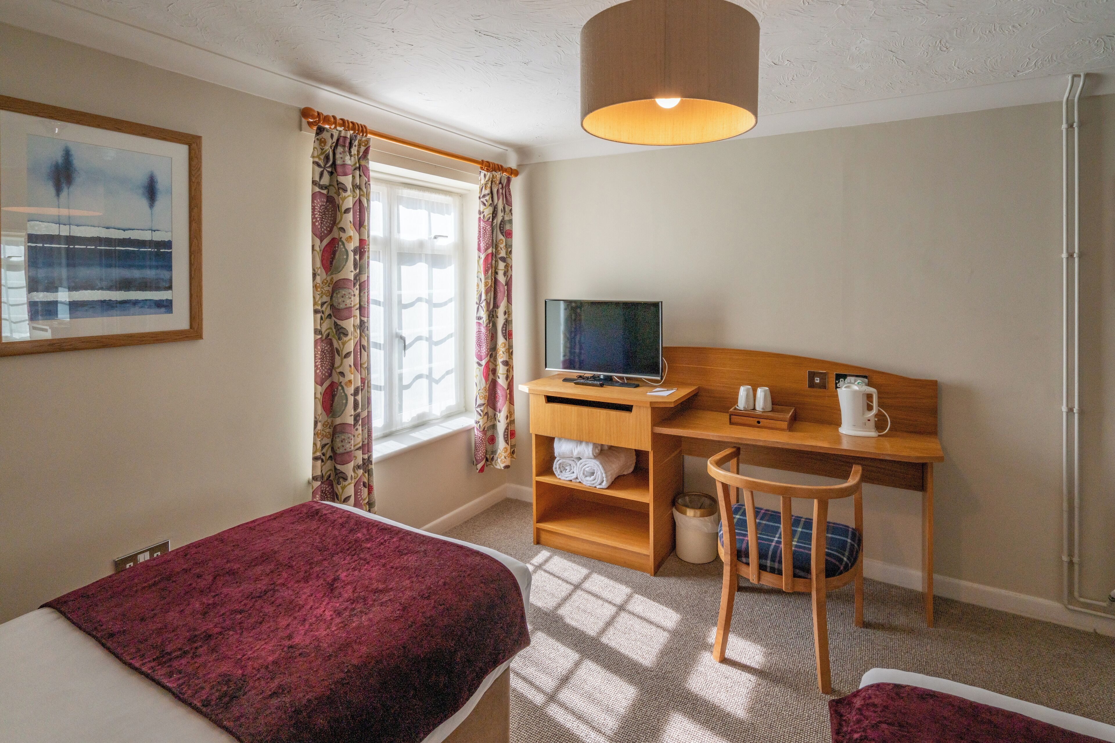 A hotel room with a bed, a desk with a computer, a chair, and a window with curtains.