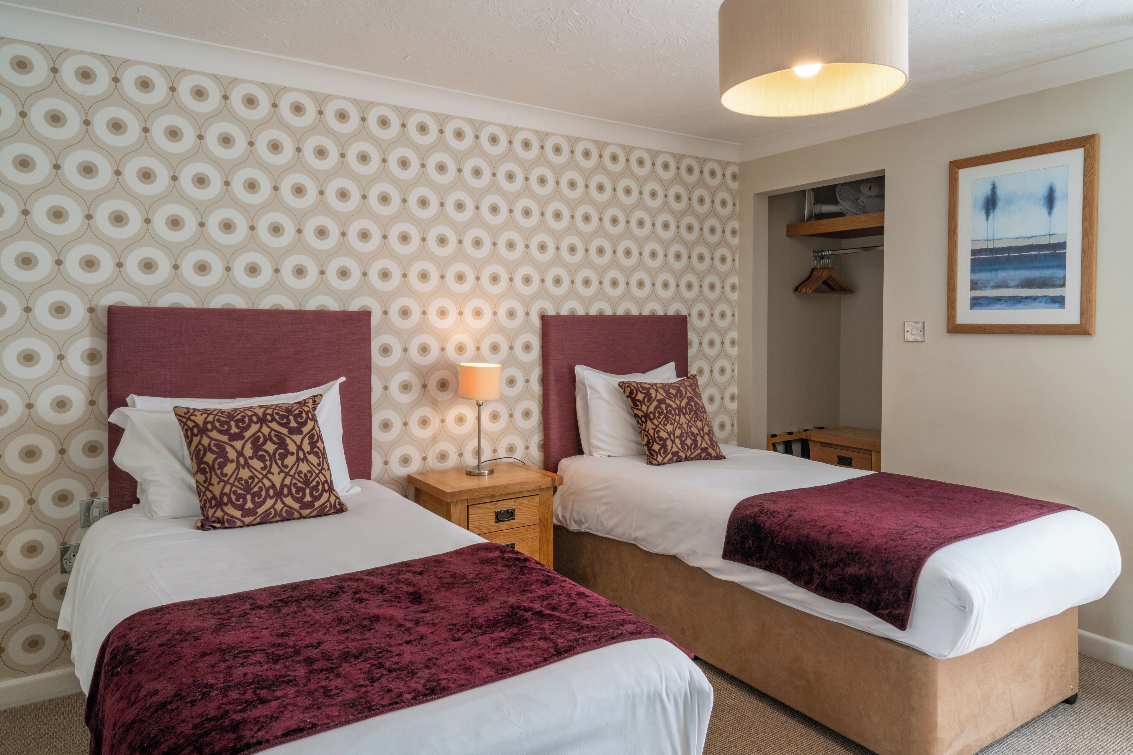 A hotel room with two beds, a patterned wallpaper, a painting, and a hanging coat rack.