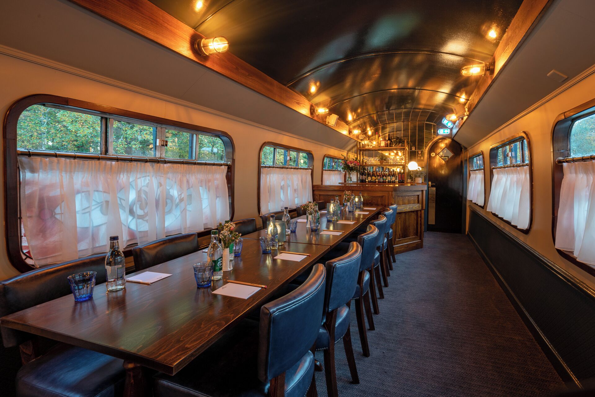 A dining car with long wooden tables, blue upholstered chairs, and a bar area with bottles and glasses.