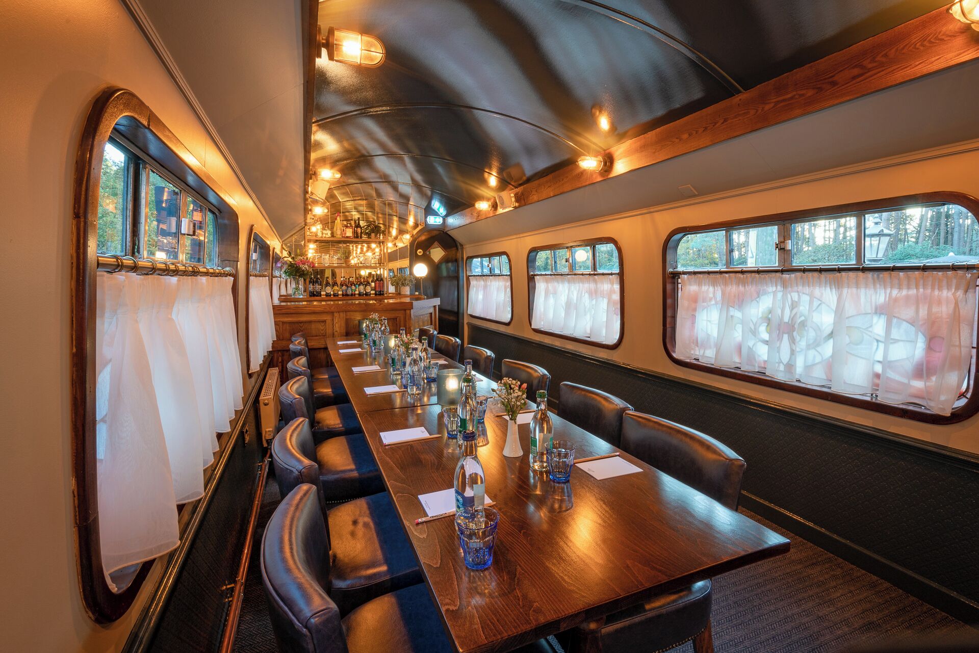 A train dining car with a long wooden table, leather seating, and large windows.