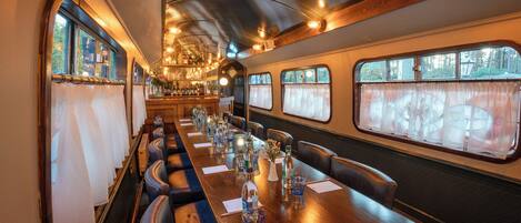 A train dining car with a long wooden table, leather seating, and large windows.