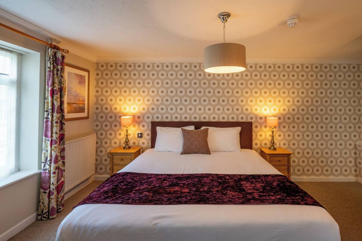 Una cama bien hecha con un edredón estampado, dos lámparas de noche y una ventana con cortinas.