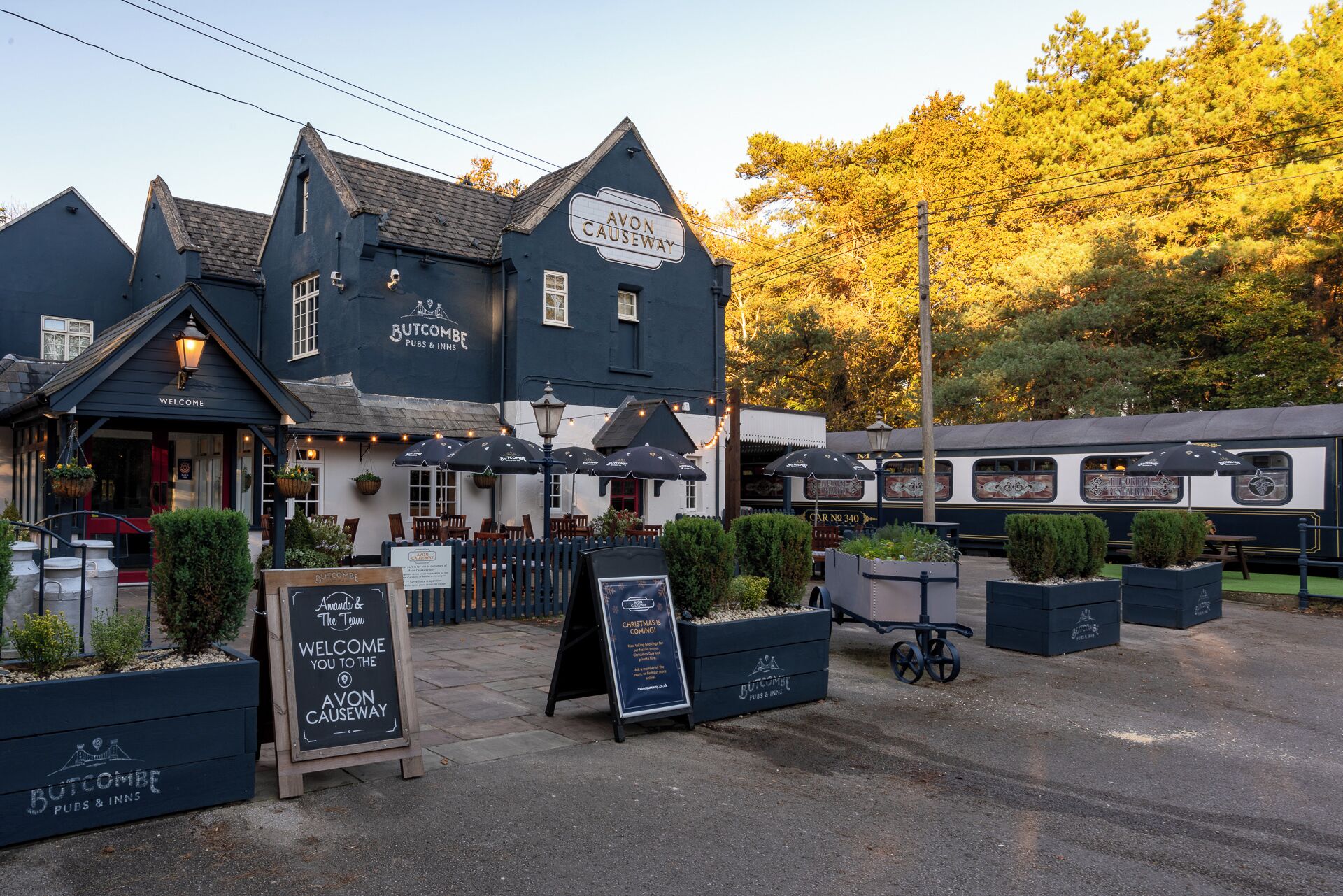 Un pub tradicional con mesas al aire libre, un letrero y un tren estacionado al fondo.