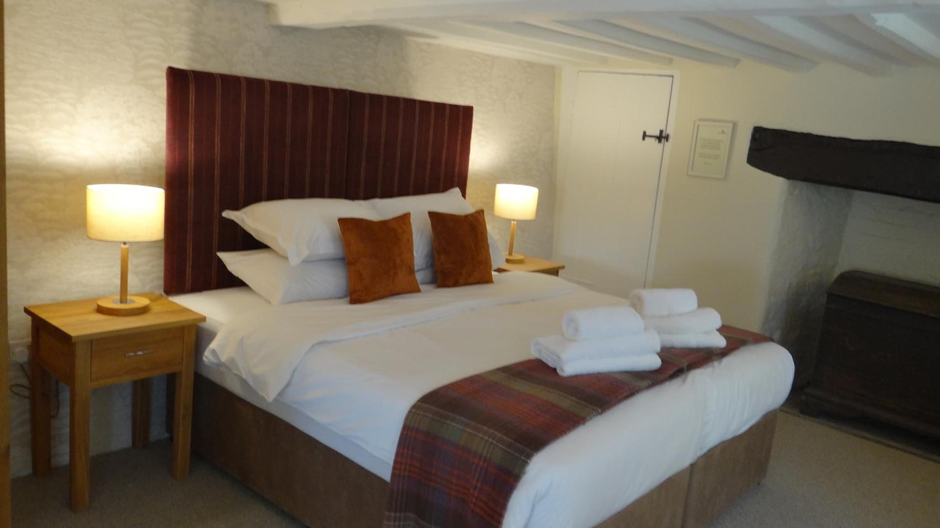 A neatly made bed with white linens and brown pillows, flanked by two bedside tables with lamps, in a room with a wooden headboard and a door in the background.