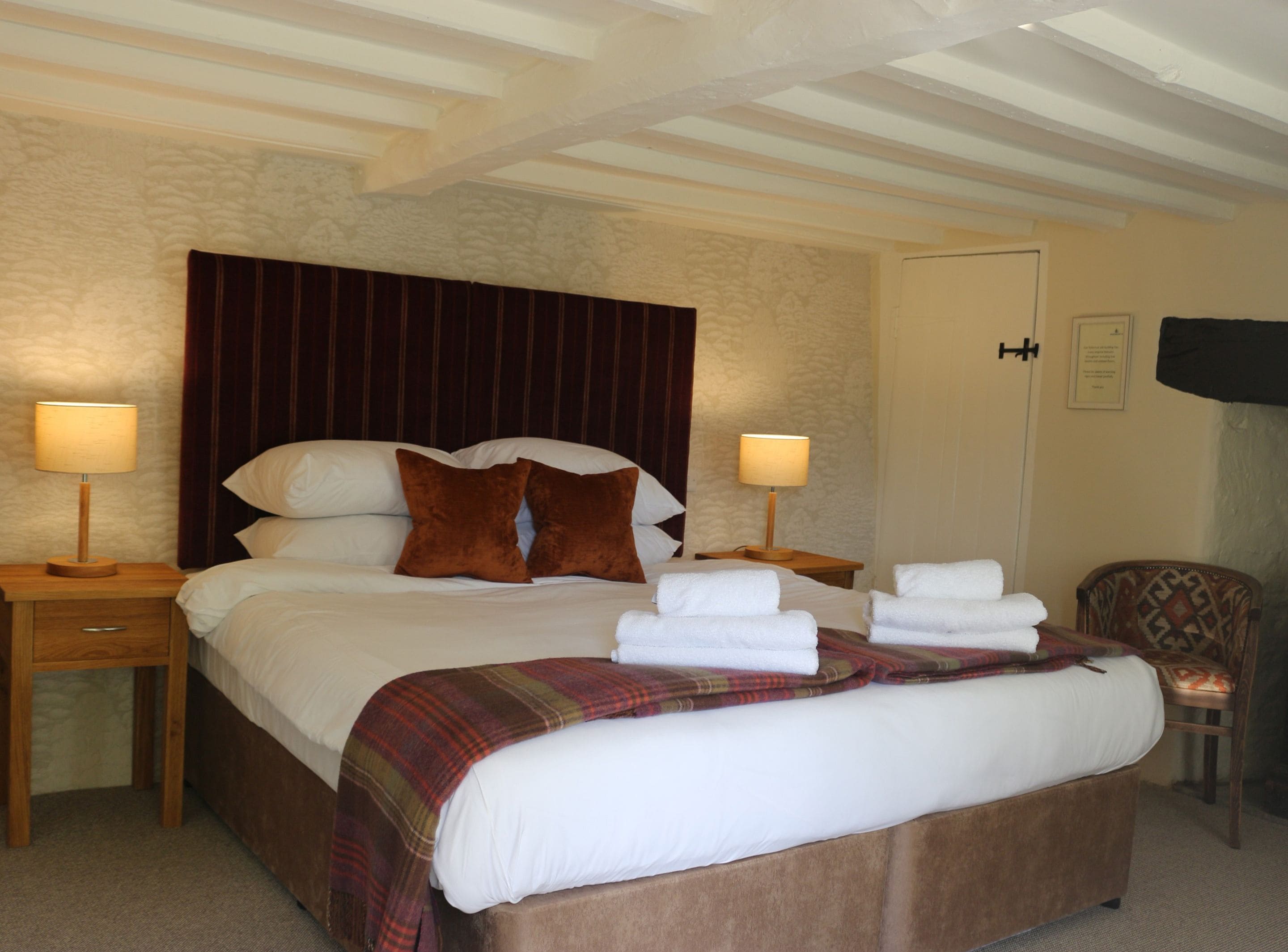 A neatly made bed with white linens, two brown pillows, and a plaid blanket. There are two bedside tables with lamps, a wooden chair, and a television on the wall.
