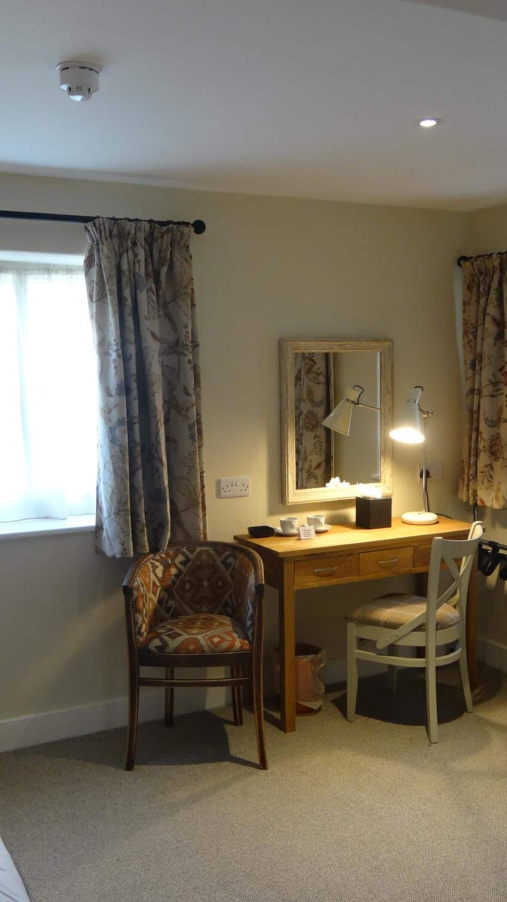 Een kamer met een houten bureau, een stoel, een bureaulamp, een spiegel en een raam met gordijnen.