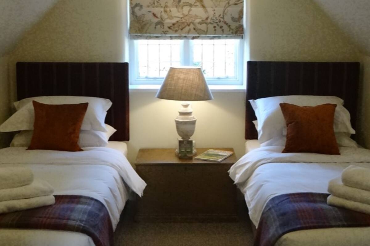 Habitación con dos camas, una ventana con estampado floral, una lámpara colgante y una mesita de noche con lámpara.