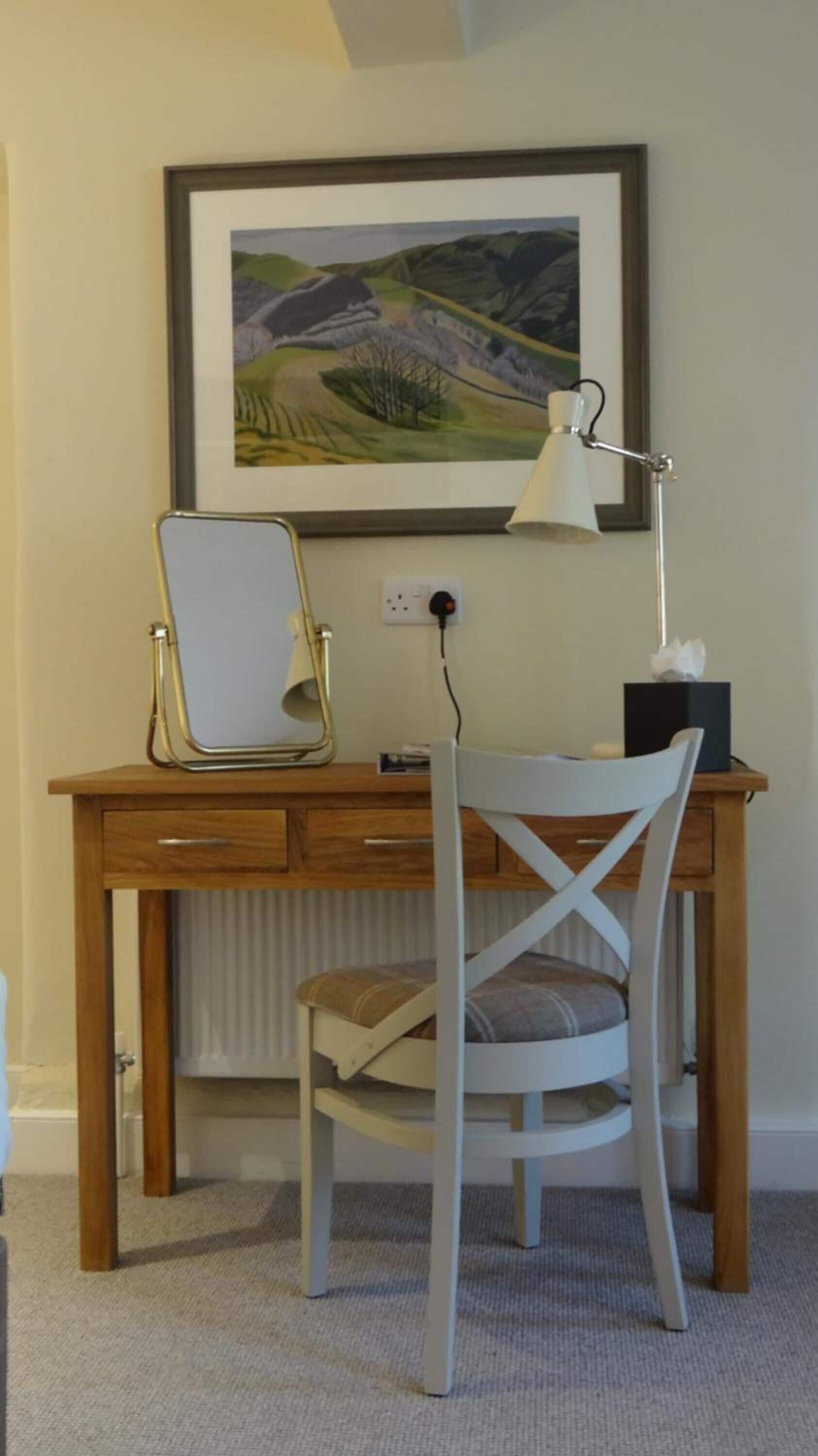A study area with a wooden desk, a white chair, a framed landscape painting, and a desk lamp.
