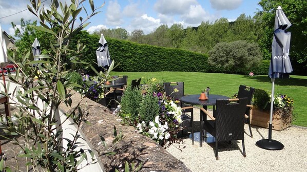 An outdoor seating area with tables and chairs, surrounded by greenery and a hedge.