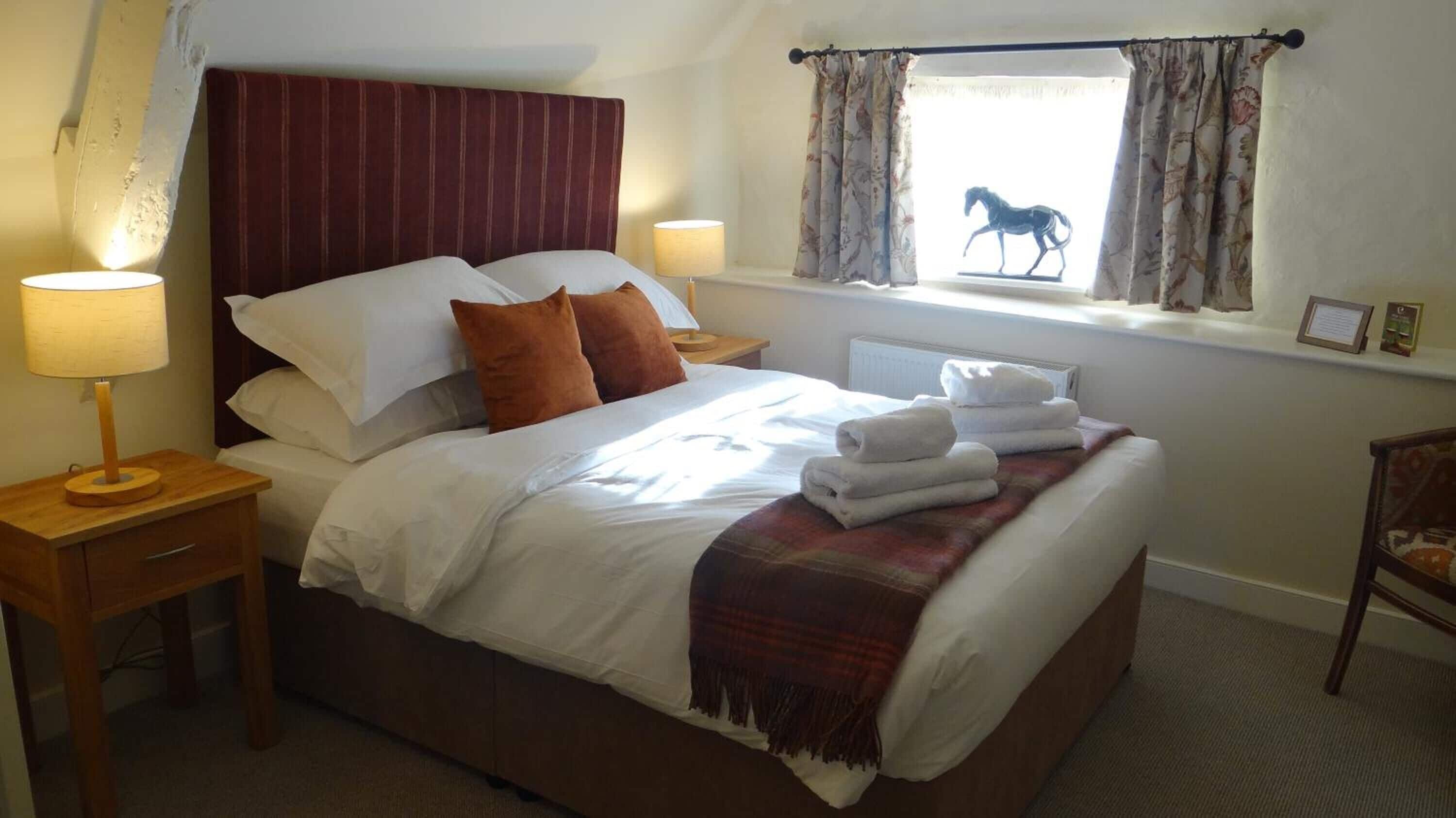 A neatly made bed with white linens, a plaid throw blanket, and folded towels on the side. There are two bedside lamps, a wooden nightstand, and a window with floral curtains and a silhouette of a horse.