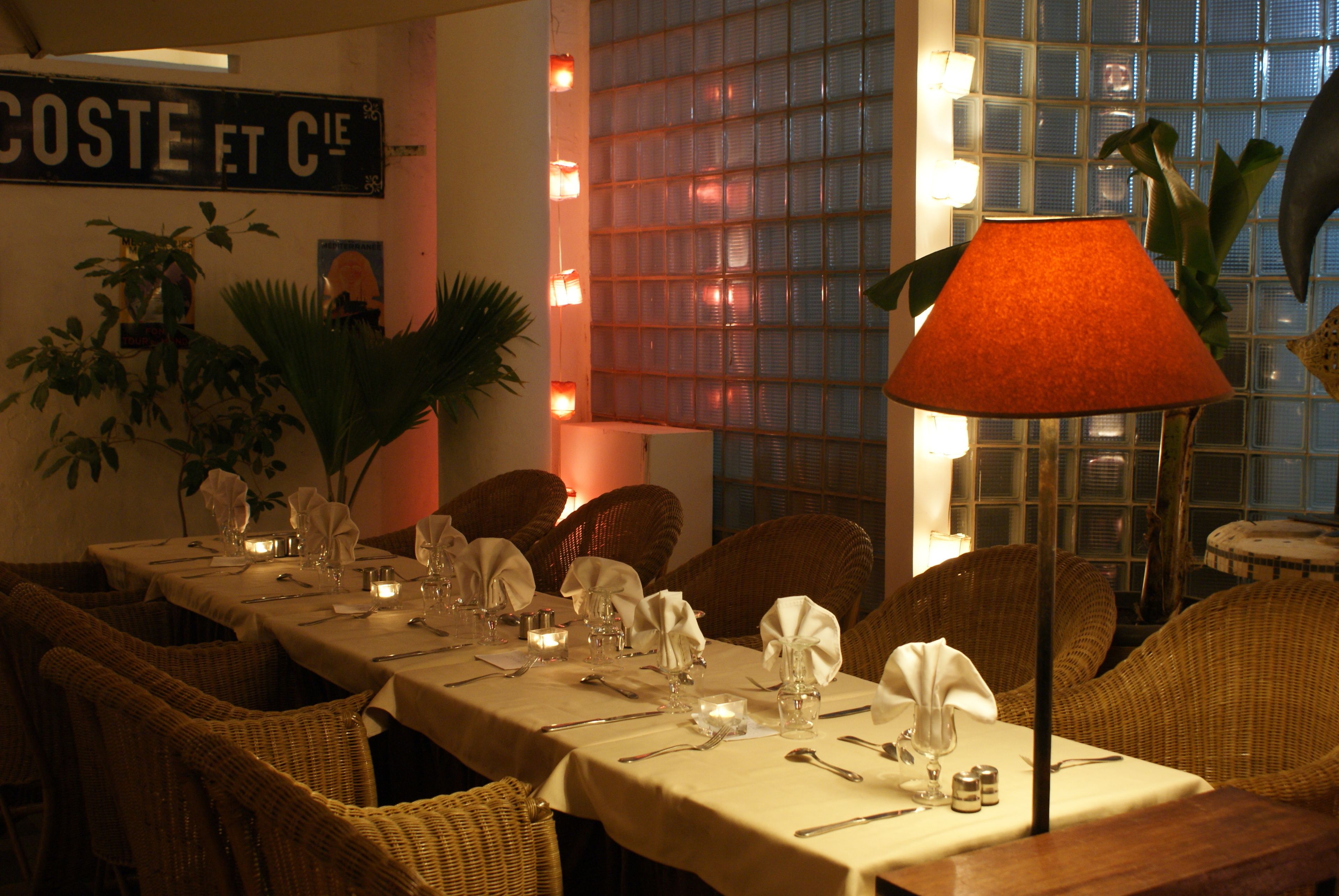 A dining table set for a meal with wicker chairs, candles, and a table lamp.