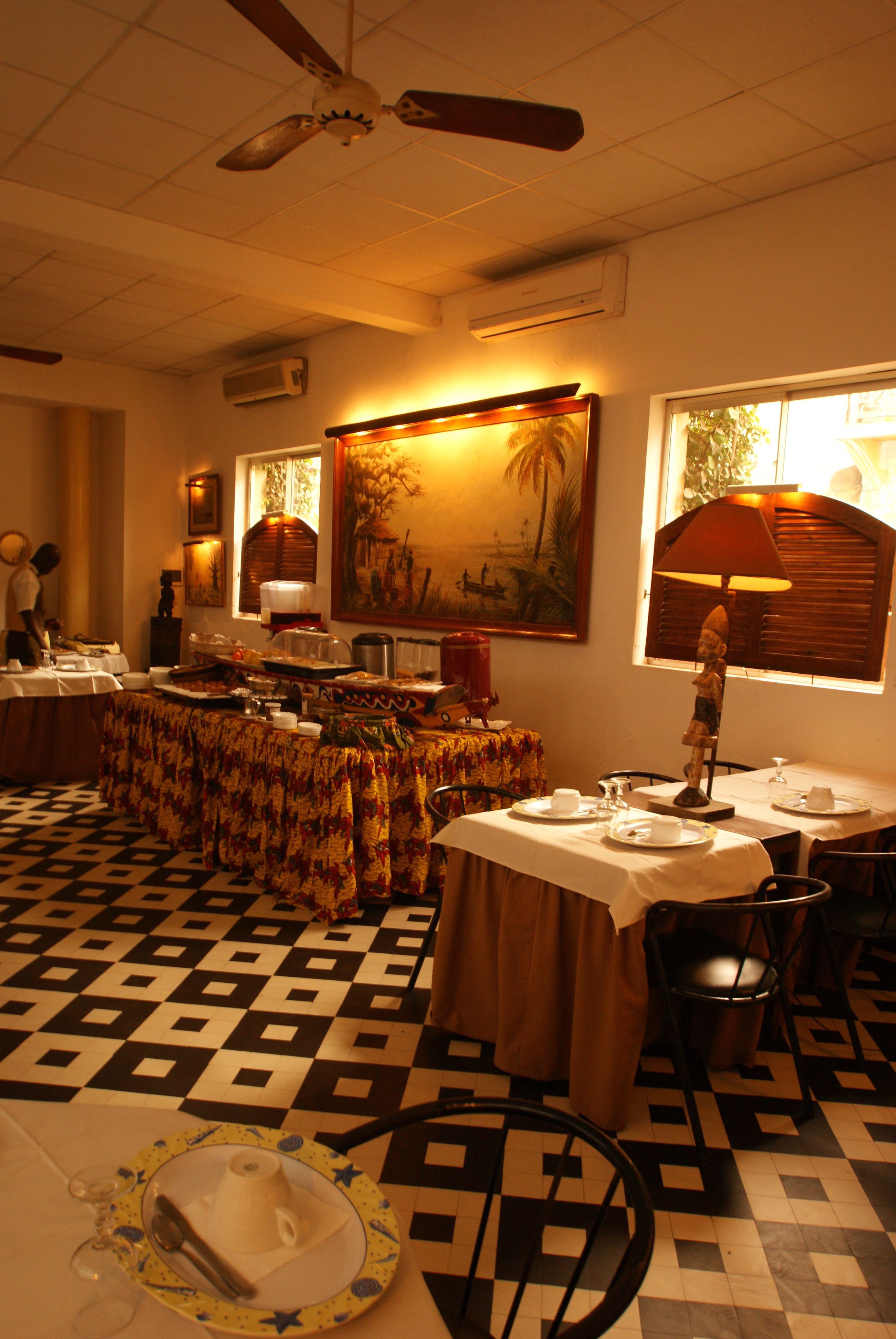 Uma sala de jantar com piso xadrez, mesas postas e um ventilador de teto.