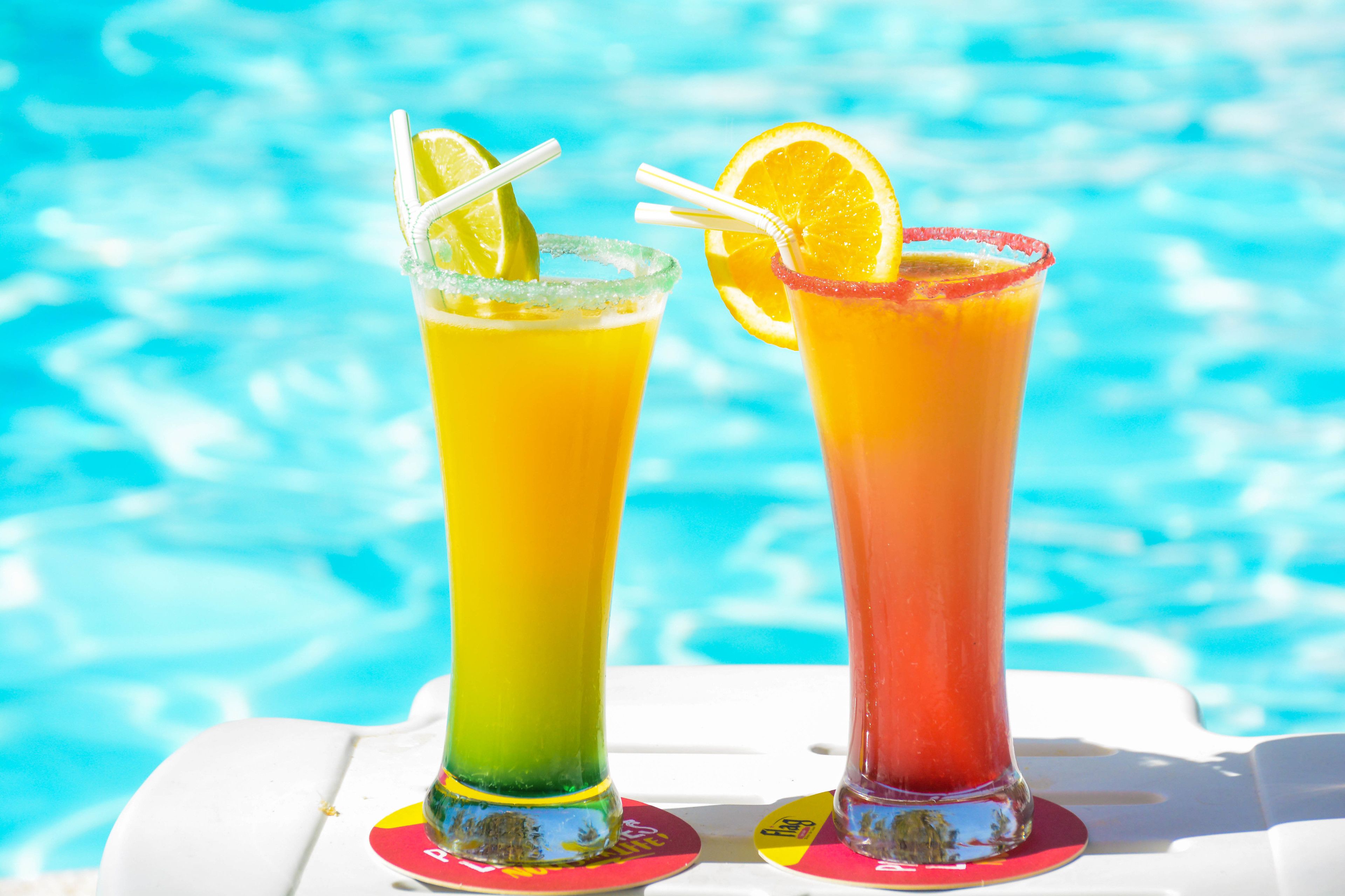 Twee cocktails met rietjes en garnering, op een witte tafel bij een zwembad.