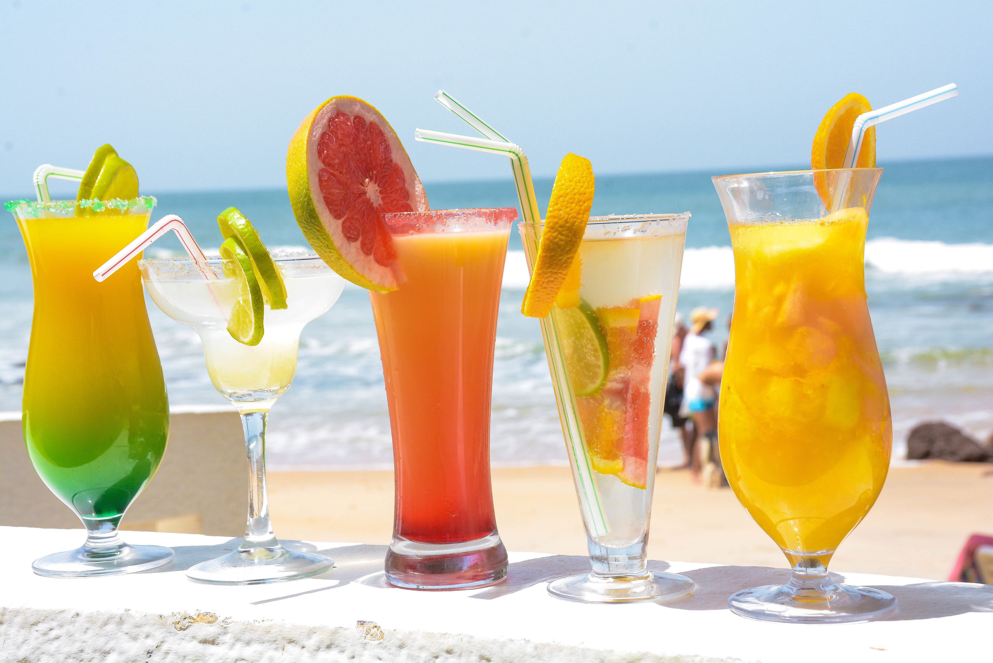 A row of colorful cocktails on a beach with the ocean in the background.