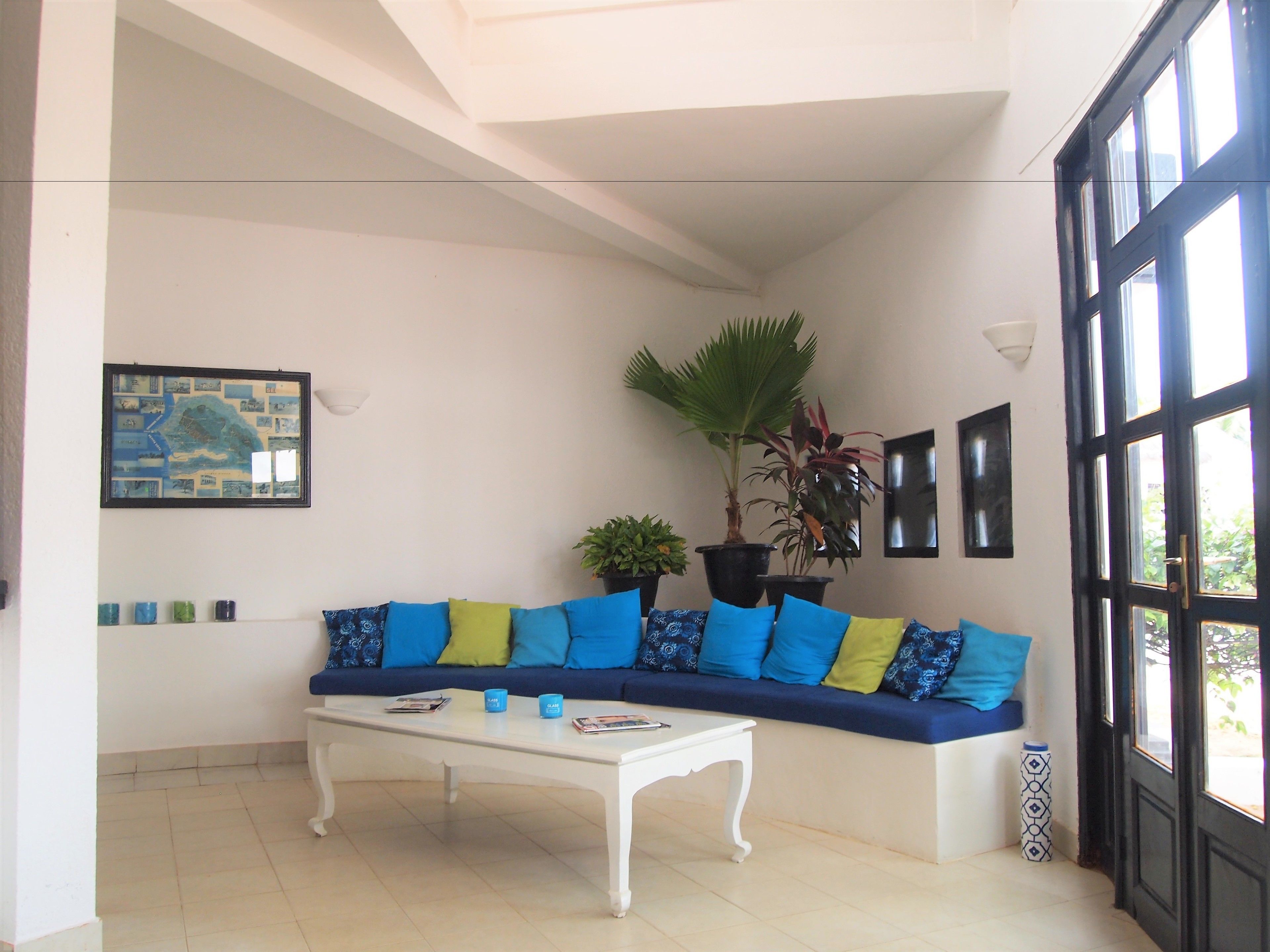Una sala de estar con un sofá blanco, cojines azules y amarillos, una mesa de centro blanca y un ventanal con marcos negros.