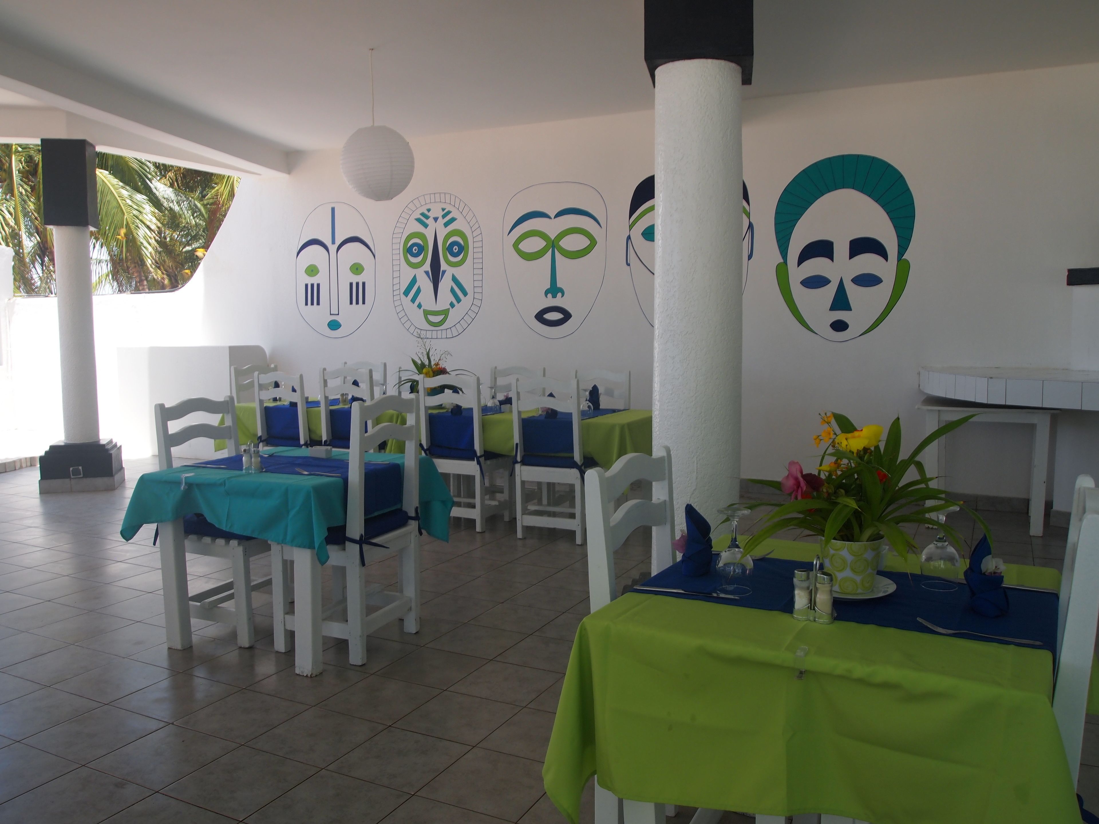 A restaurant with white chairs and tables, some with green tablecloths. There are decorative wall art pieces and a white pillar.