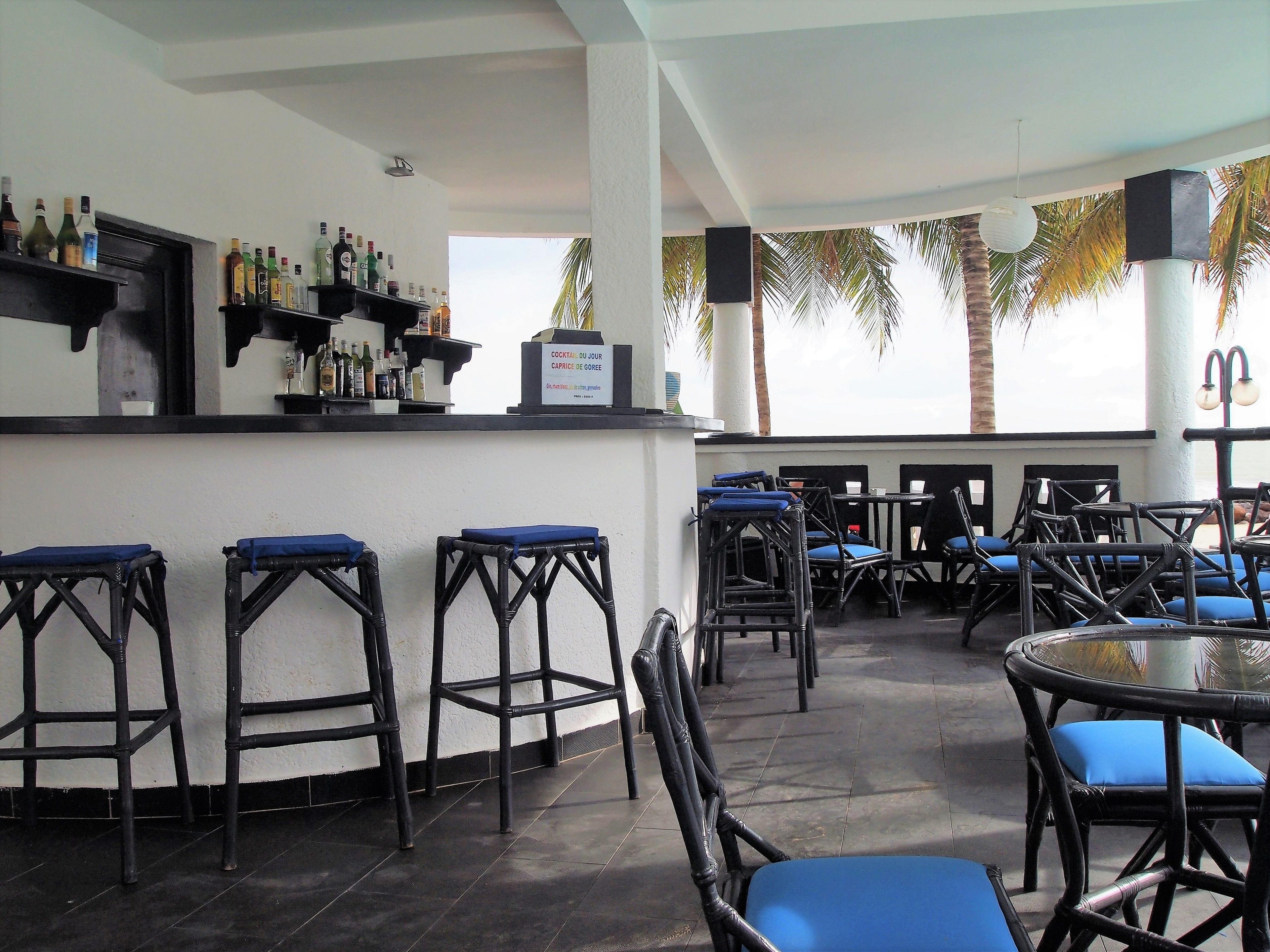 A bar area with stools and tables, a sign indicating a cocktail menu, and palm trees in the background.
