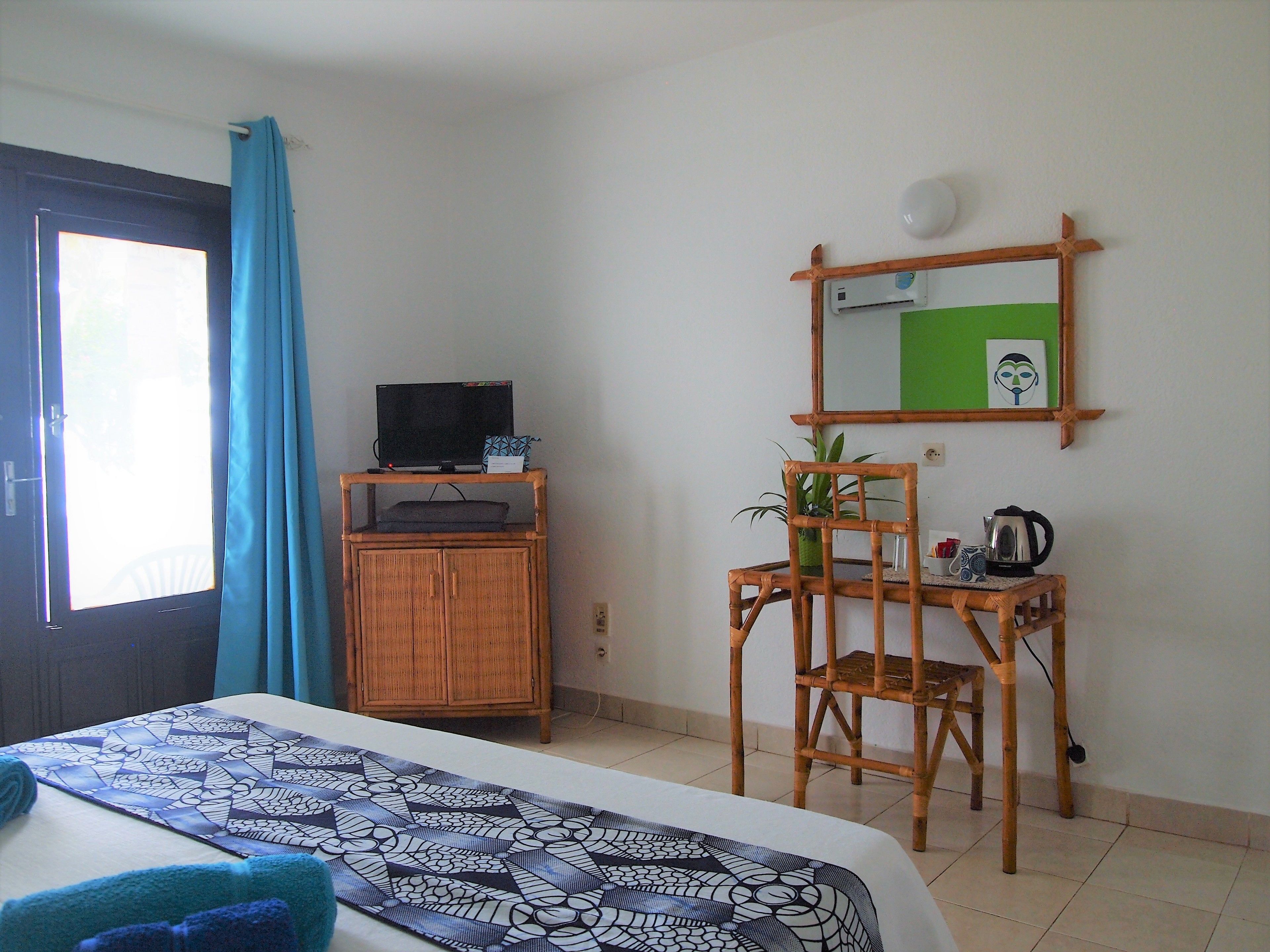 A room with a bed, a television on a wooden cabinet, a chair, a table with a kettle, and a mirror on the wall.
