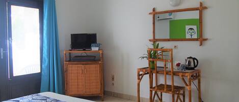 A room with a bed, a television on a wooden cabinet, a chair, a table with a kettle, and a mirror on the wall.