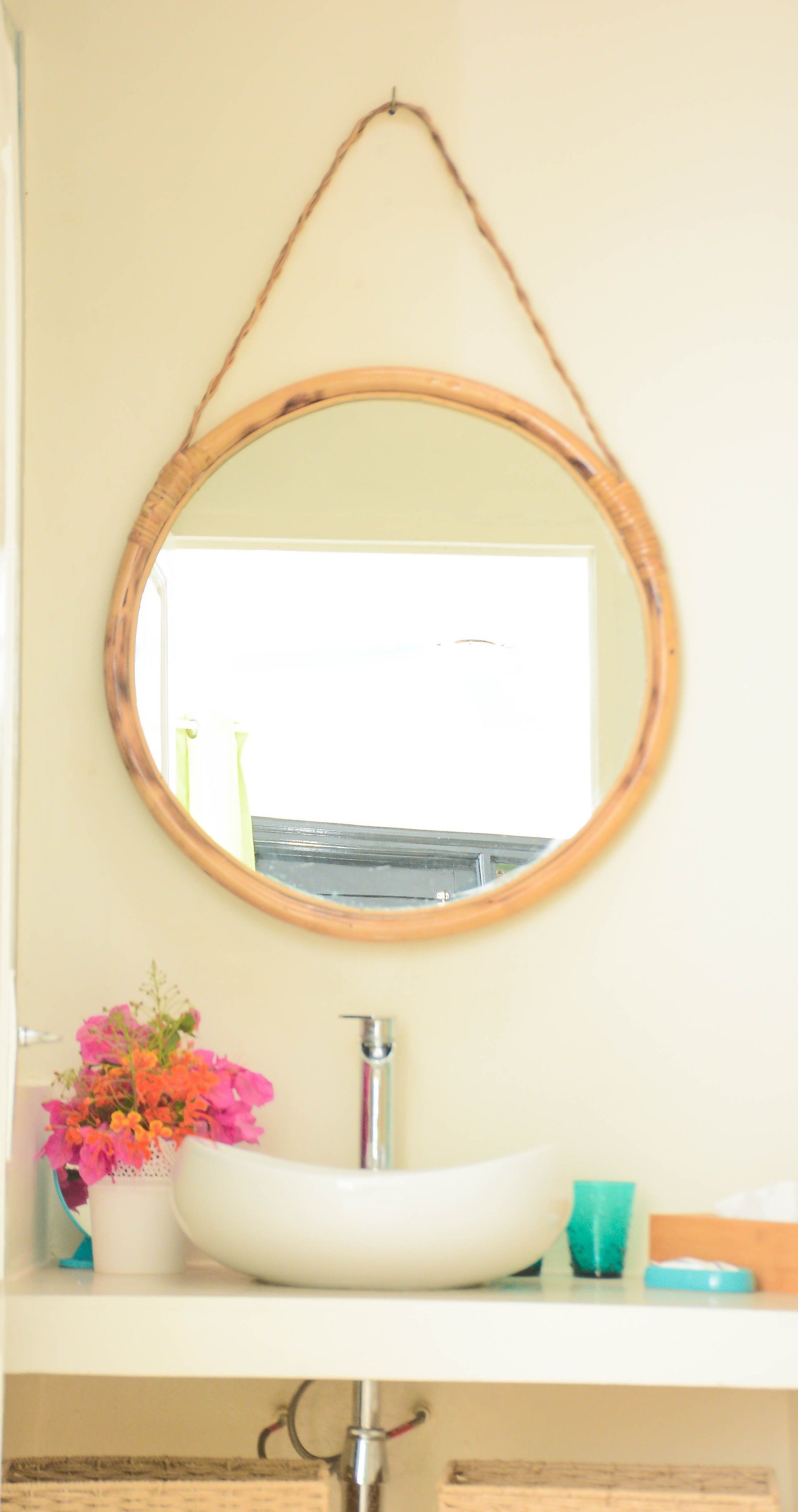 A bathroom with a round mirror, a white sink, a green cup, and a vase with pink flowers.