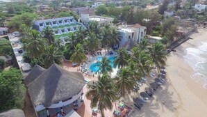 Un resort con piscina central rodeada de palmeras, asientos frente a la playa y un edificio con techo de paja.
