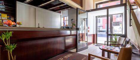 A reception area with a wooden counter, a reception desk, a glass door leading outside, and a seating area with a table and chairs.