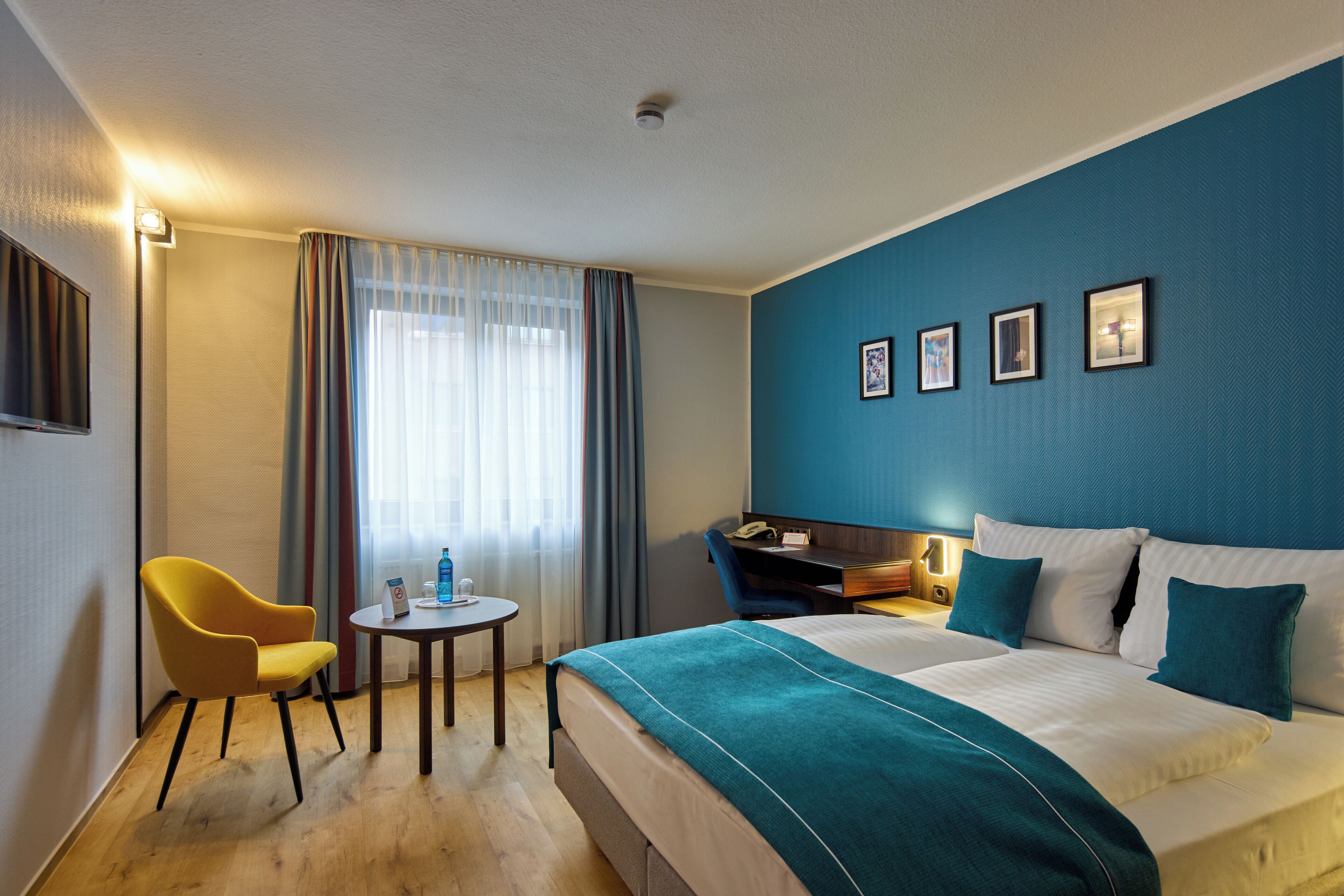 A hotel room with a bed, a desk, a chair, a small table, and a window with curtains.