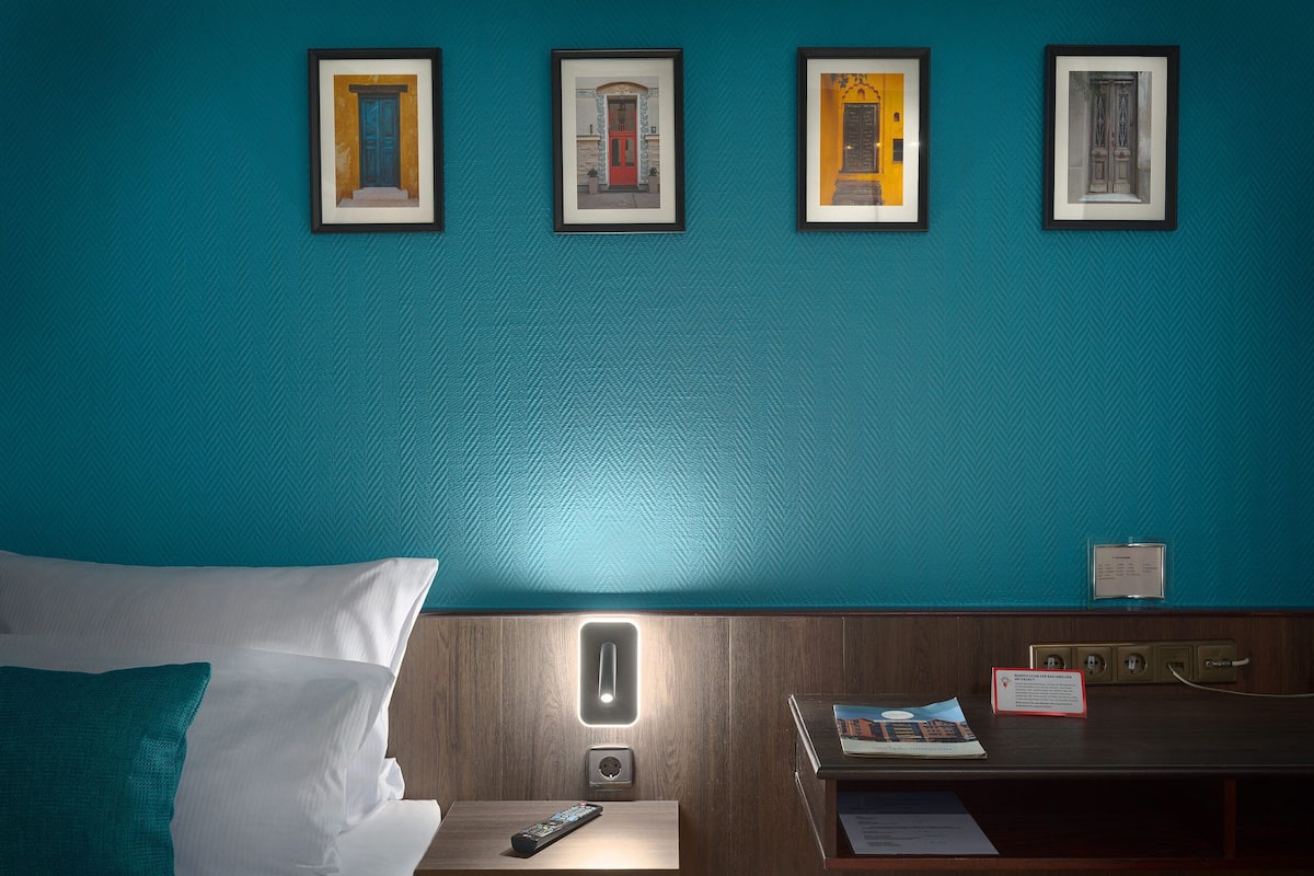 Een hotelkamer met teal muren, een bed met witte en teal kussens, een nachtkastje met een afstandsbediening, en ingelijste foto’s aan de muur.