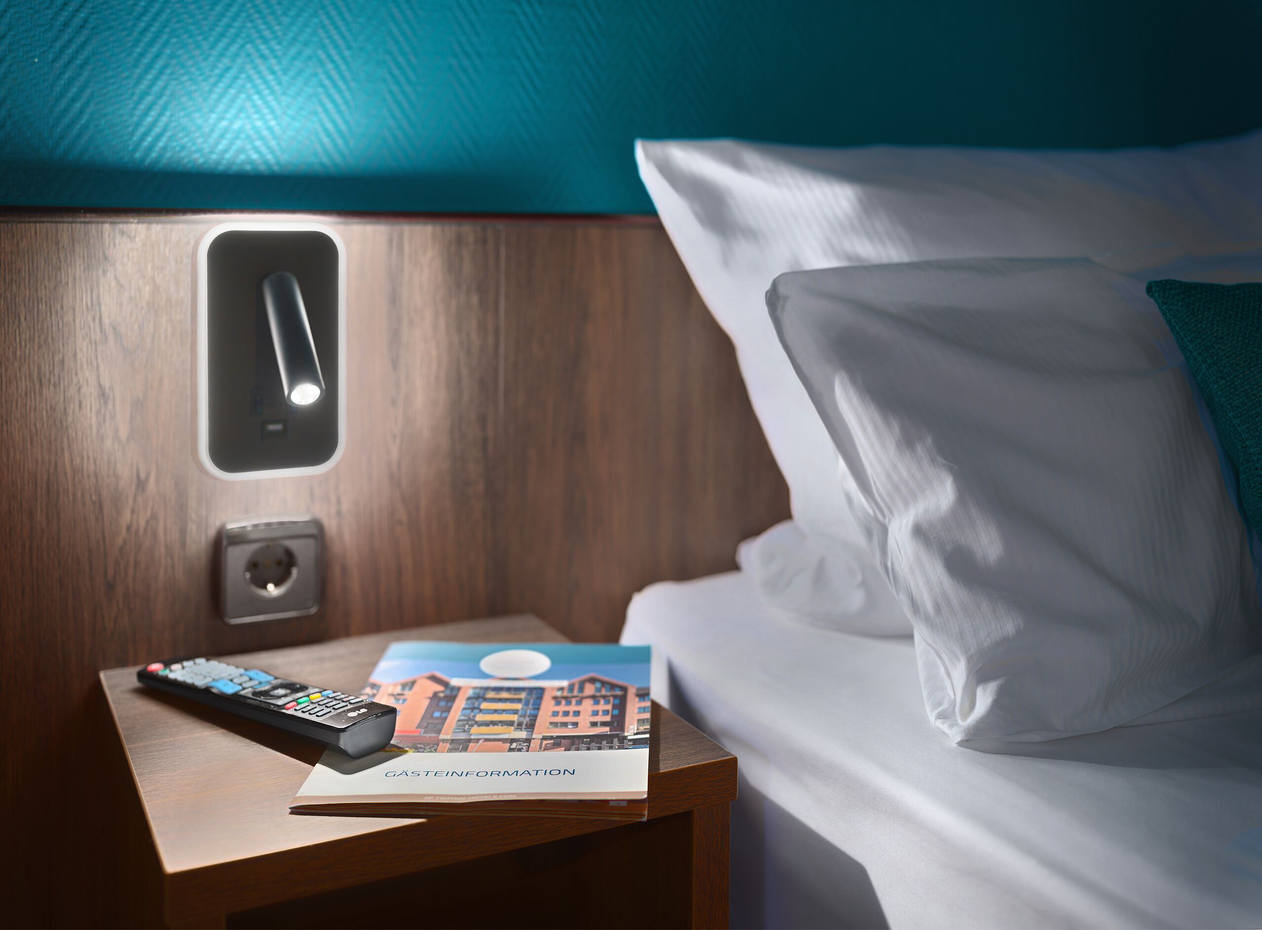 Une chambre d’hôtel avec une table de chevet en bois, une télécommande et une brochure intitulée « GASTEINFORMATION ».