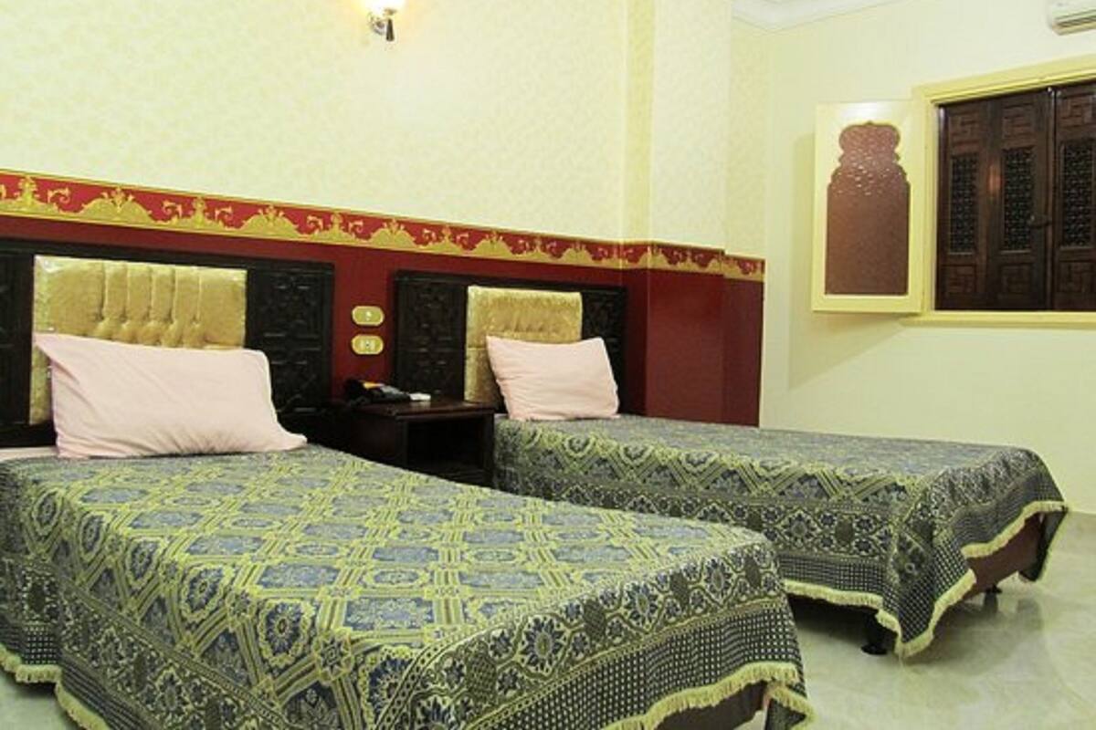 A hotel room with two beds, each with a patterned bedspread, a red and gold headboard, and a wall-mounted light fixture.