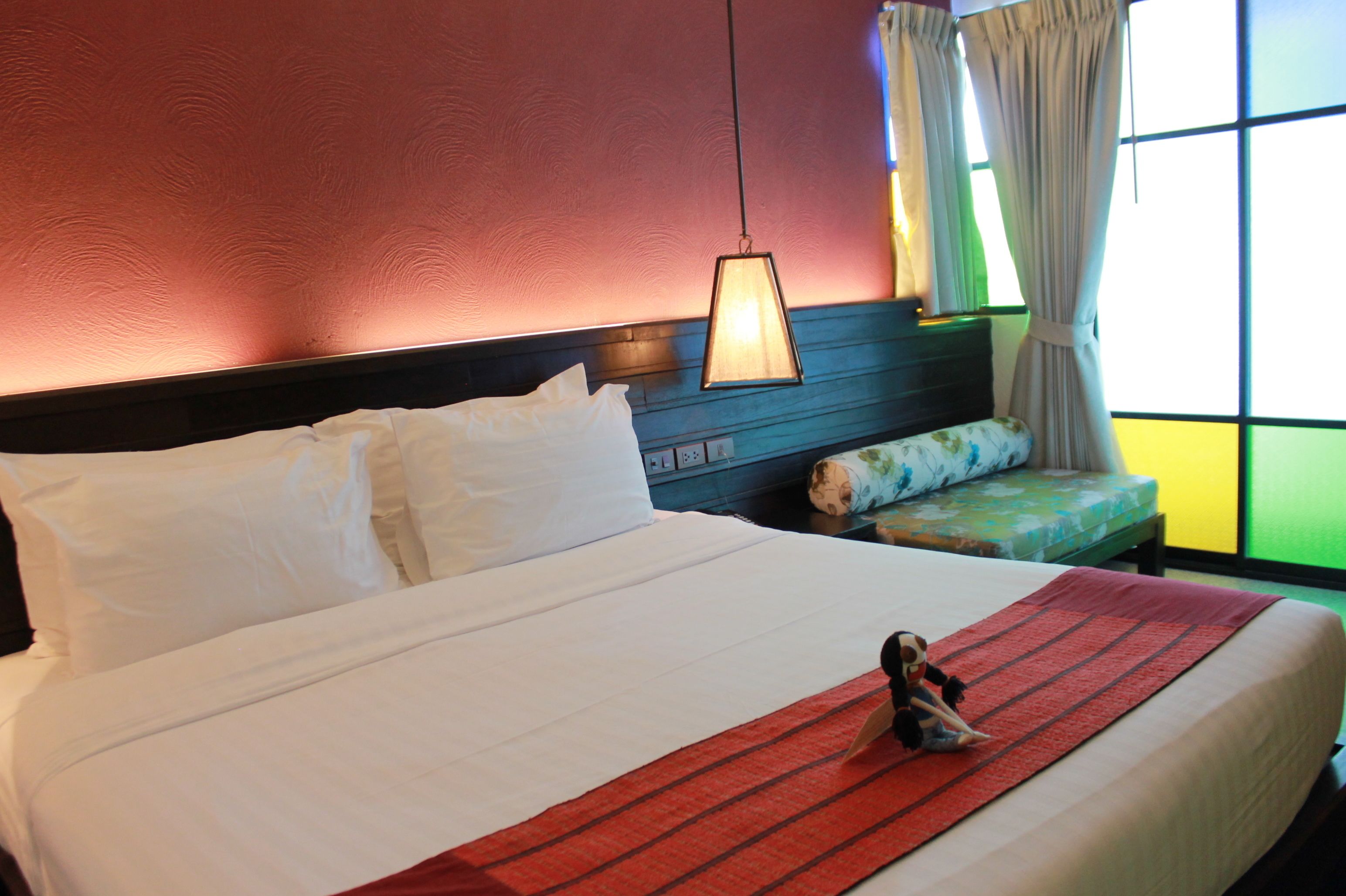 A hotel room with a bed, pillows, a bedside lamp, a window with curtains, and a small bench with a floral cushion.