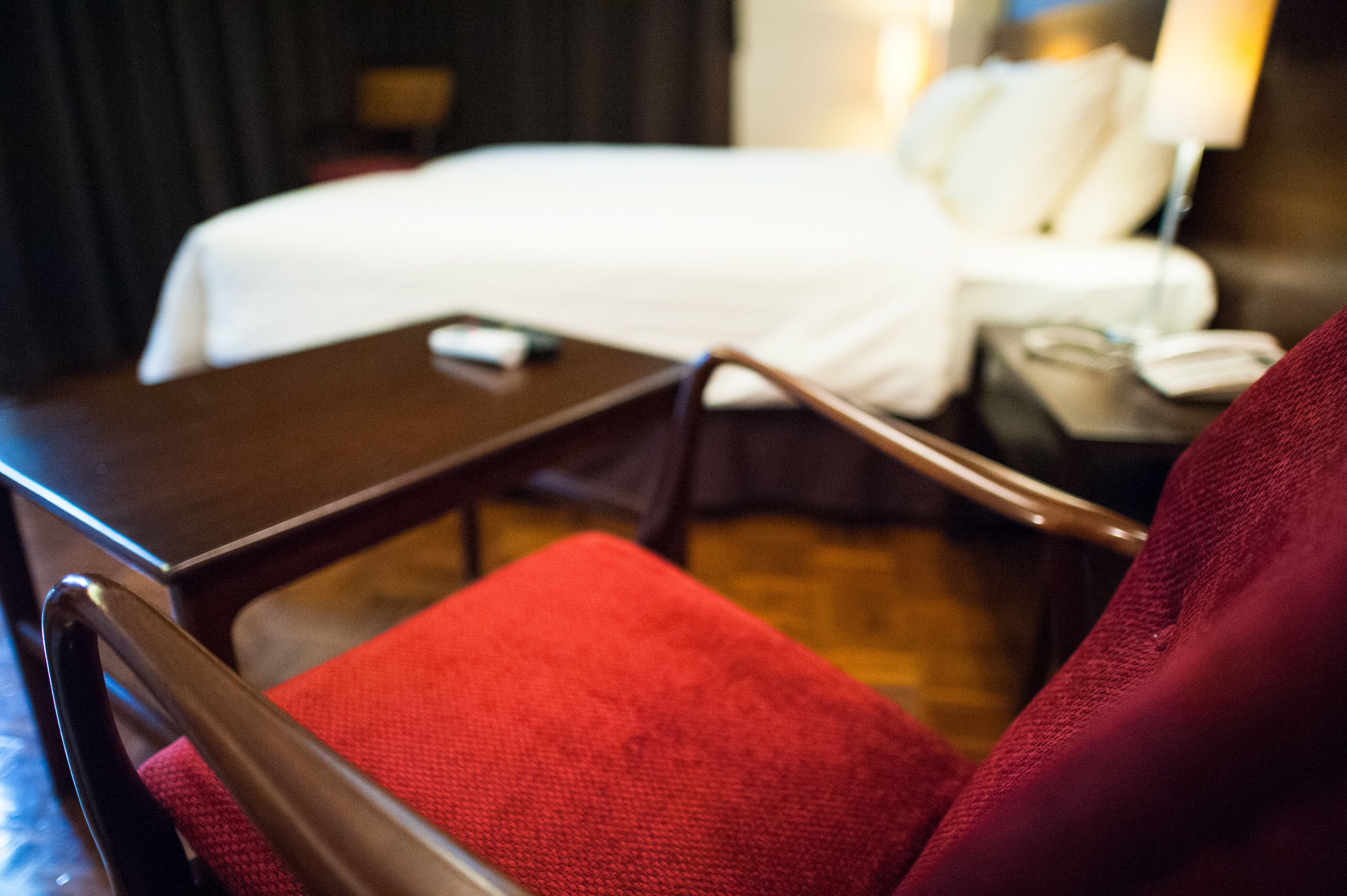 A hotel room with a bed, a bedside table, a chair, and a red cushion.