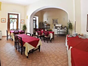 Comedor con mesas cubiertas con manteles rojos, zona de cocina y un amplio arco.