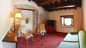 A cozy living room with a stone archway, a wooden ceiling, a sofa, two striped chairs, a small table with a lamp, and a television. - Hotel Della Fonte (Greccio)