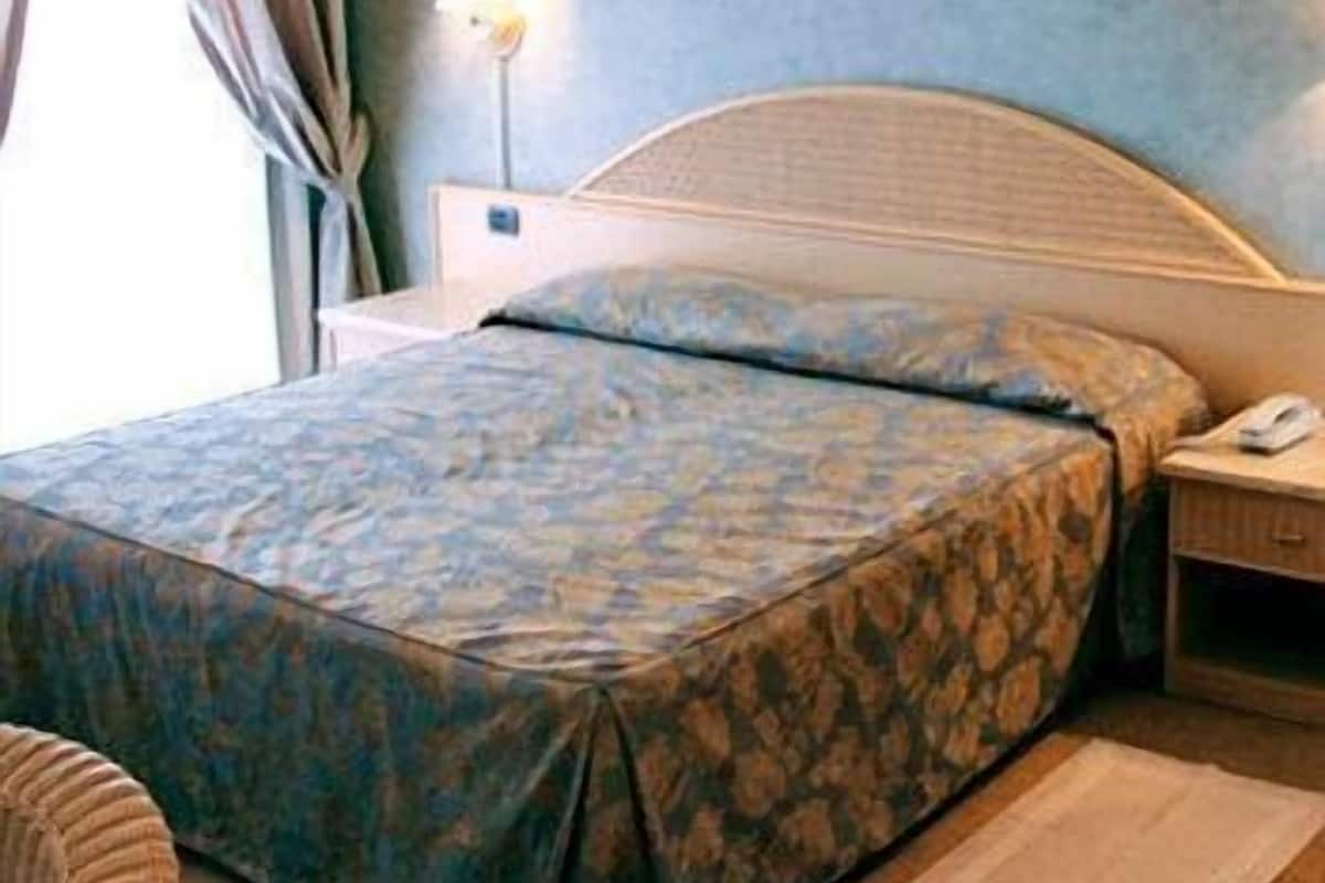 Quarto com cama, mesa de cabeceira, abajur e janela com cortinas.