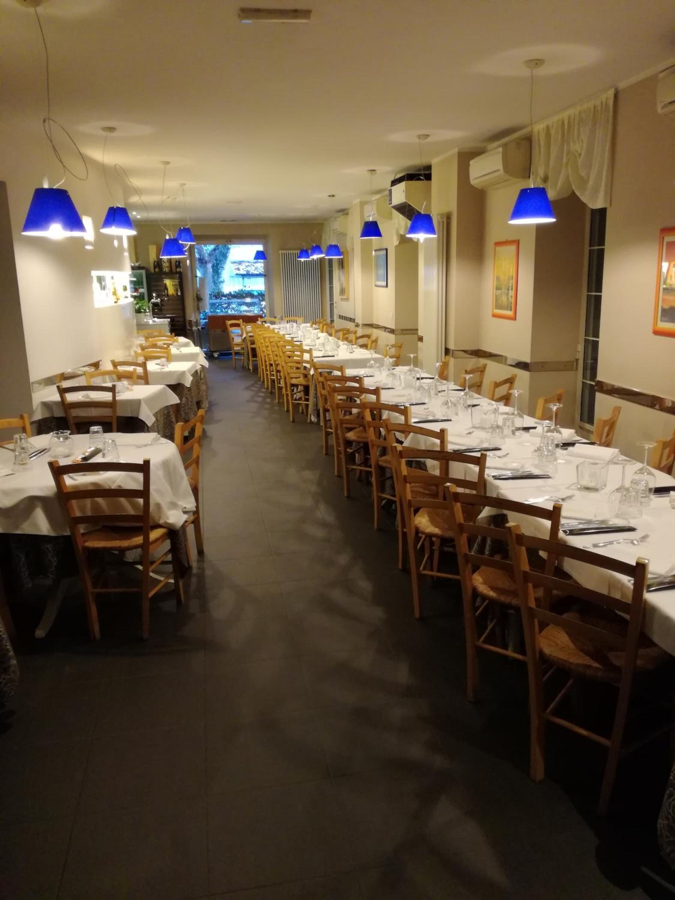 A long dining hall with multiple tables set for a meal, wooden chairs, and blue pendant lights.