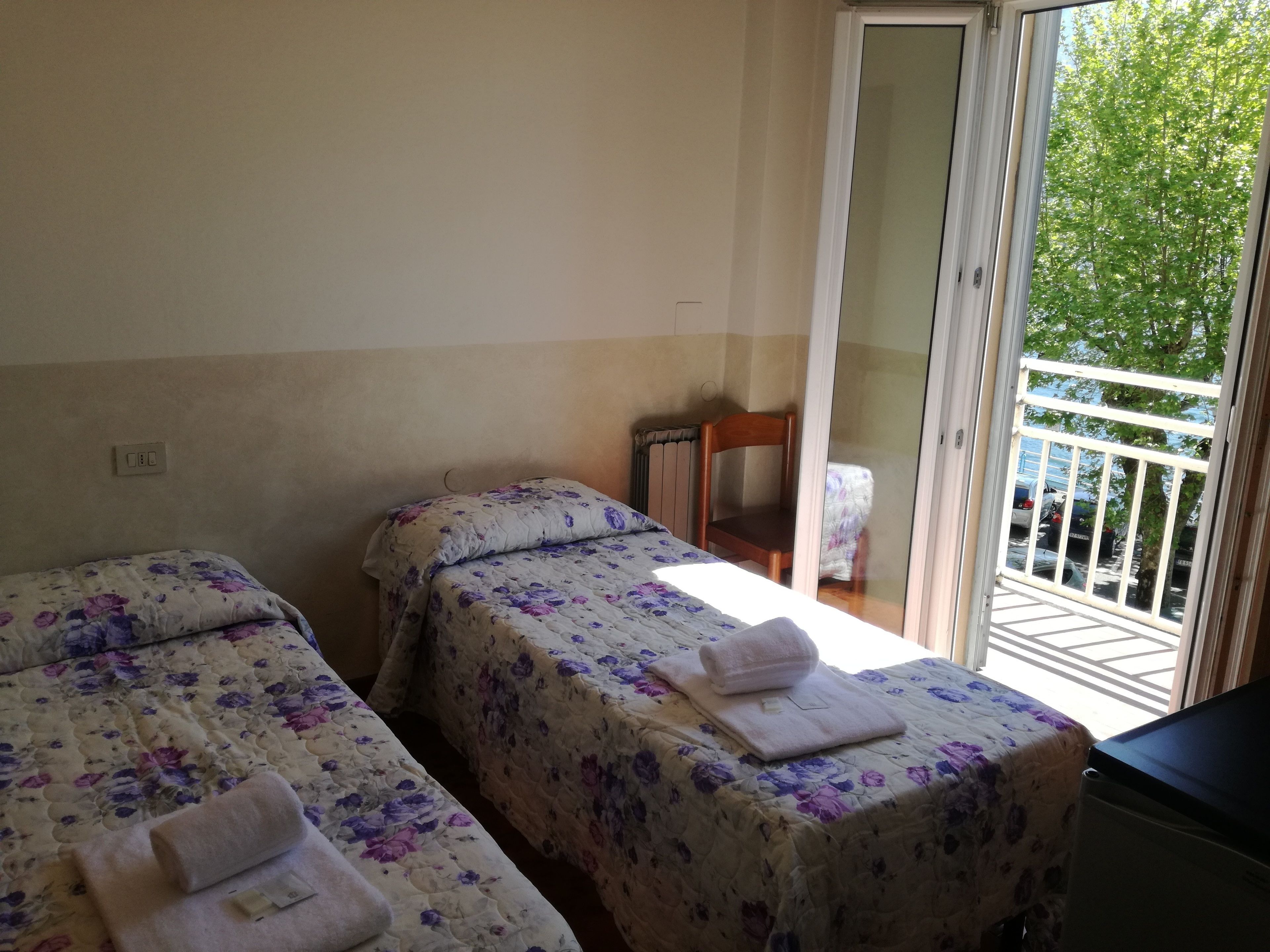 Ein Zimmer mit zwei Betten, jeweils mit blumigem Bettzeug und Handtüchern ausgestattet. Ein Sessel und eine Heizung sind an der Wand zu sehen. Eine Glastür öffnet sich zu einem Außenbereich mit Blick auf parkende Autos und Bäume.