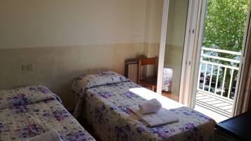 Ein Zimmer mit zwei Betten, jeweils mit blumigem Bettzeug und Handtüchern ausgestattet. Ein Sessel und eine Heizung sind an der Wand zu sehen. Eine Glastür öffnet sich zu einem Außenbereich mit Blick auf parkende Autos und Bäume.