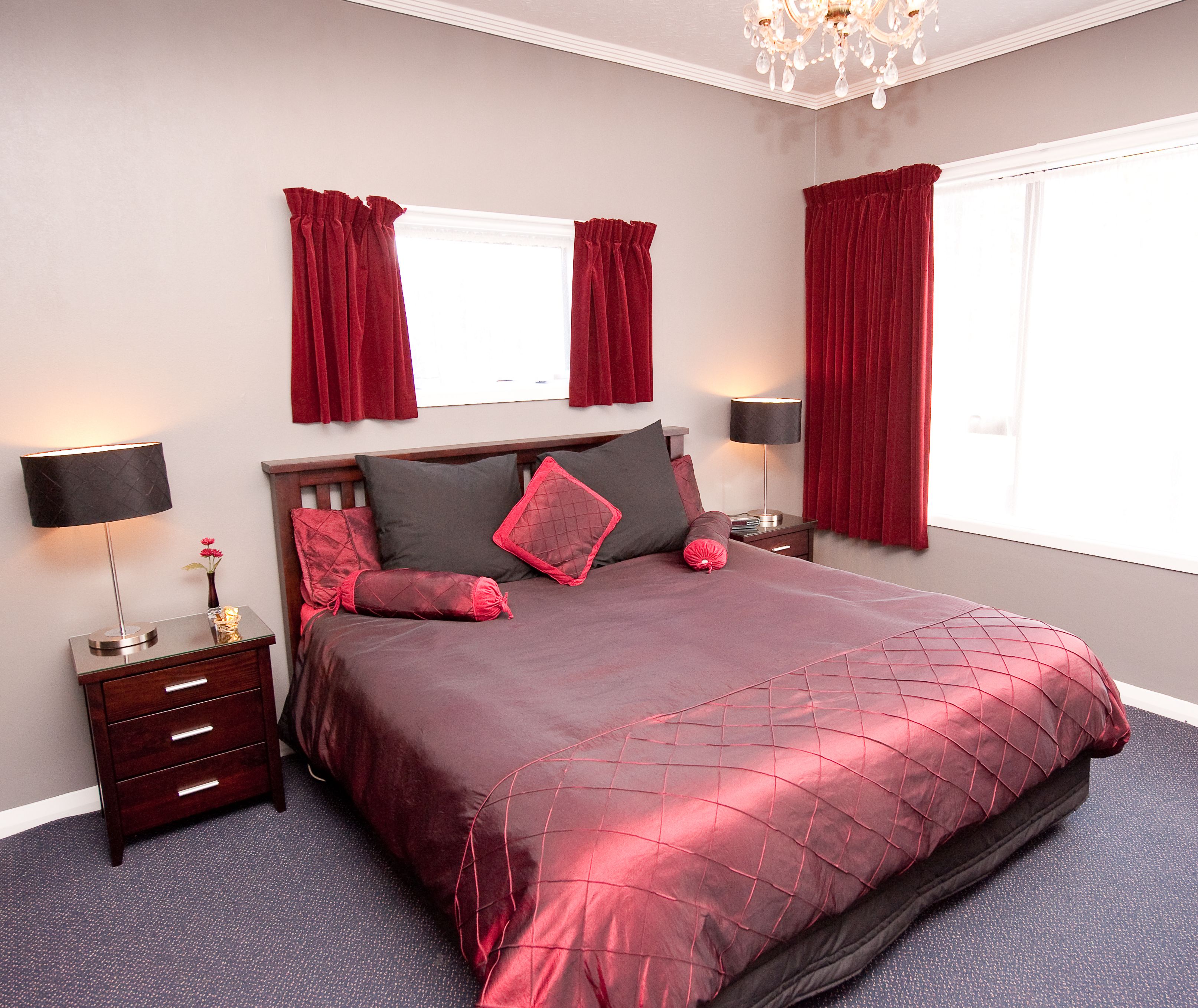 A neatly made bed with a red bedspread, two bedside tables with lamps, a dresser, and a chandelier.