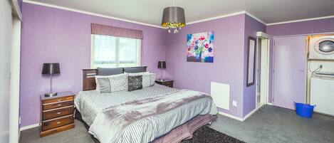 Une chambre à coucher avec un lit, des tables de chevet, une commode, une fenêtre avec des rideaux, un tableau, une porte avec un lave-linge et un seau bleu.
