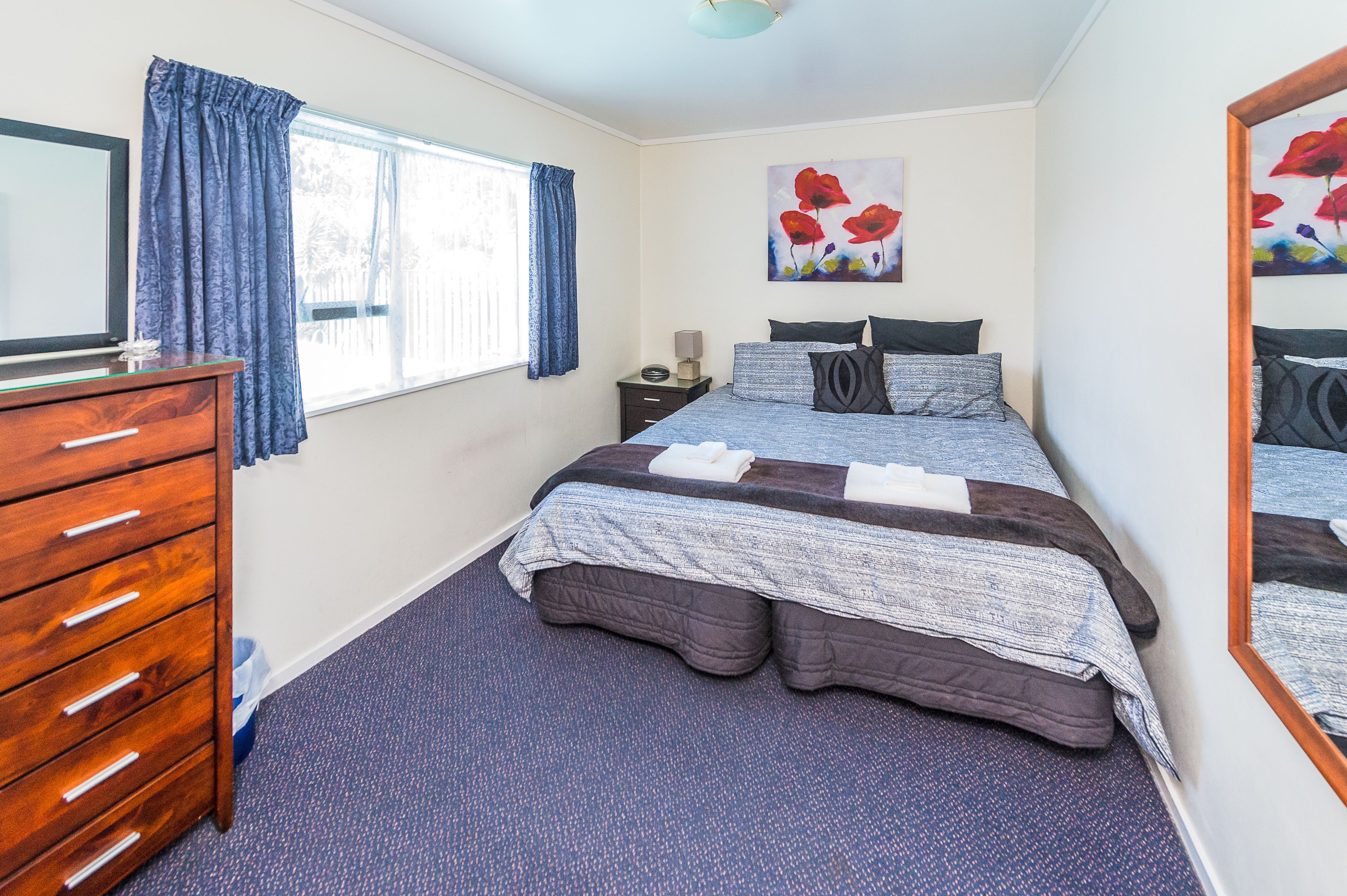 A bedroom with a bed, dresser, window with curtains, bedside table, and artwork.