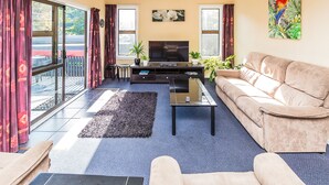A living room with a sofa, a coffee table, a TV, and a large window with curtains. - Anndion Lodge Motel and Function Centre (Whanganui)