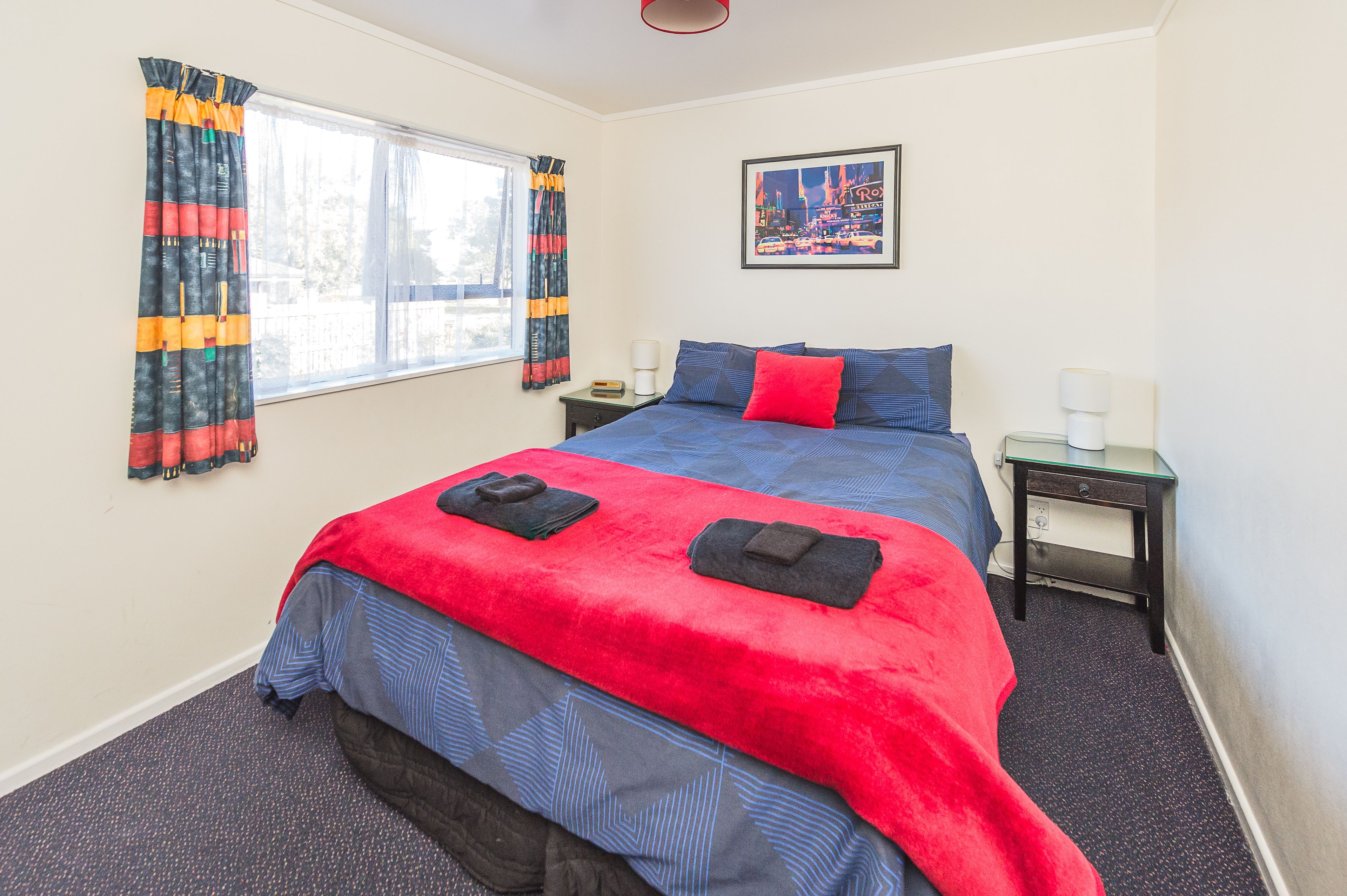A bedroom with a large bed, two bedside tables, a window with curtains, and a framed picture on the wall.