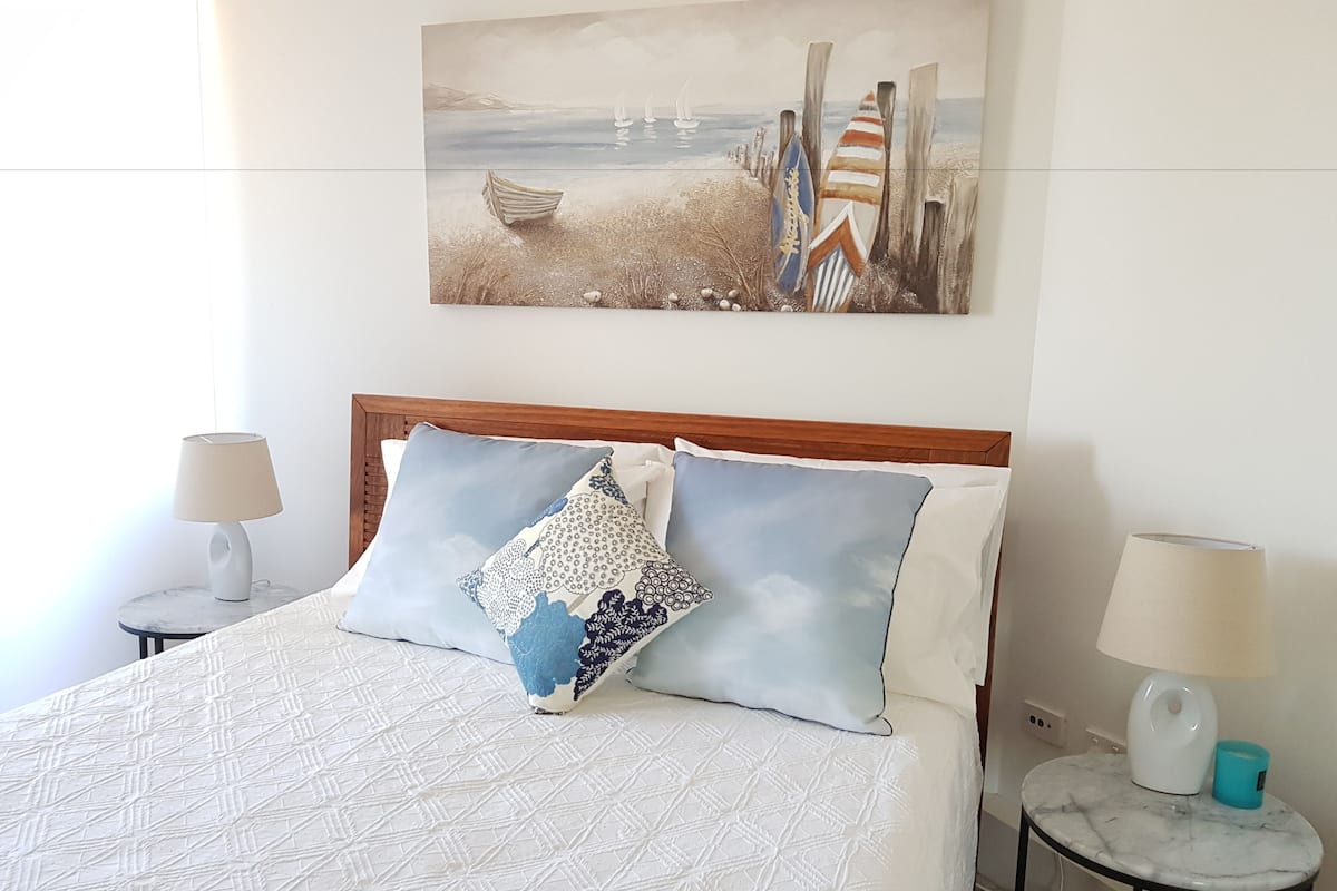 A neatly made bed with patterned bedding, two bedside tables with lamps, and a painting of a beach scene above the bed.