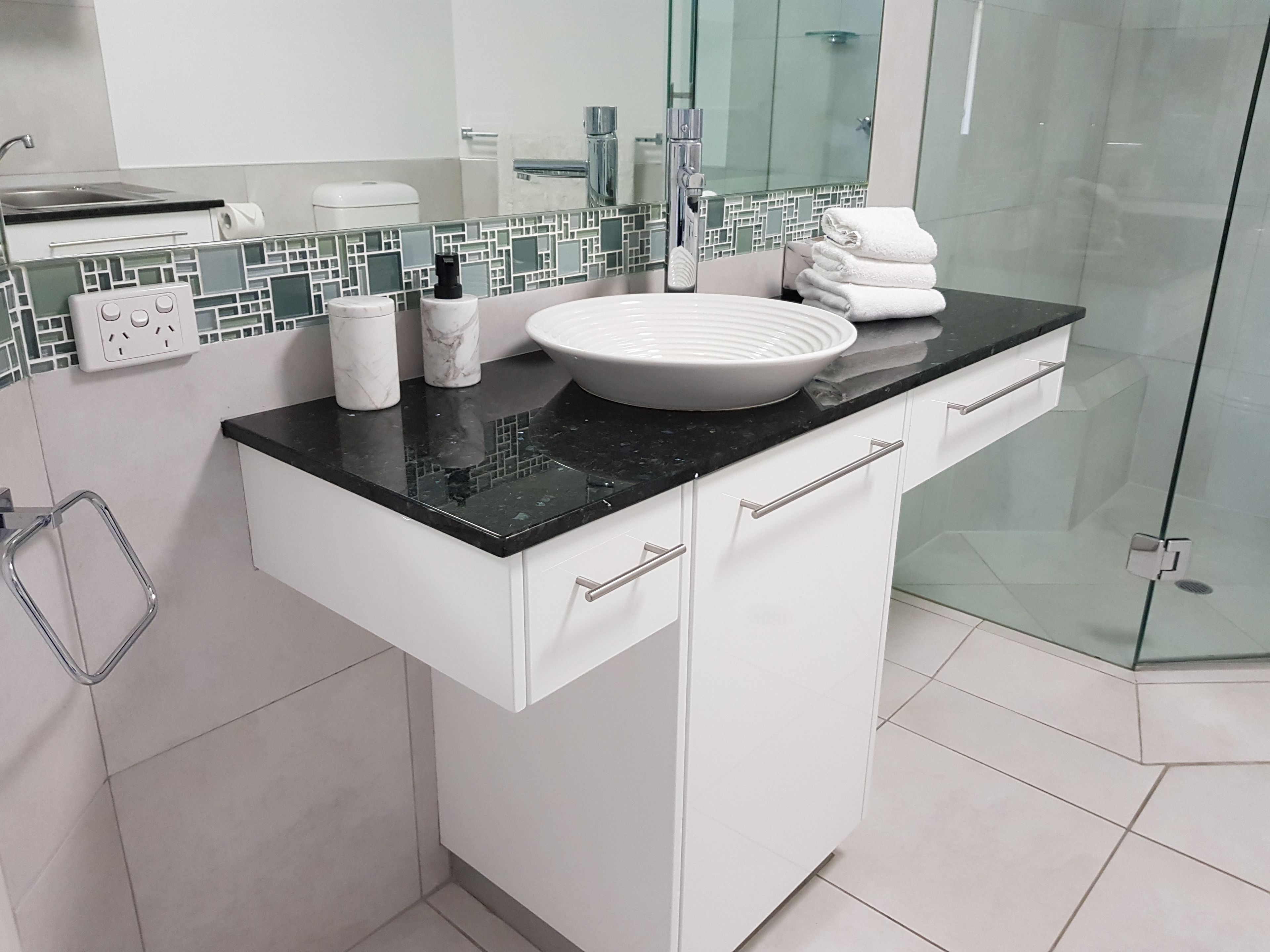 A modern bathroom with a black countertop, white cabinets, a glass shower enclosure, and a tiled backsplash.