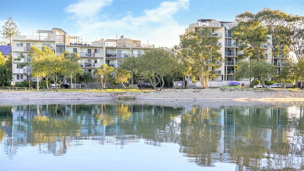 Edifício residencial com várias varandas, uma praia arenosa e uma superfície de água calma refletindo o ambiente.