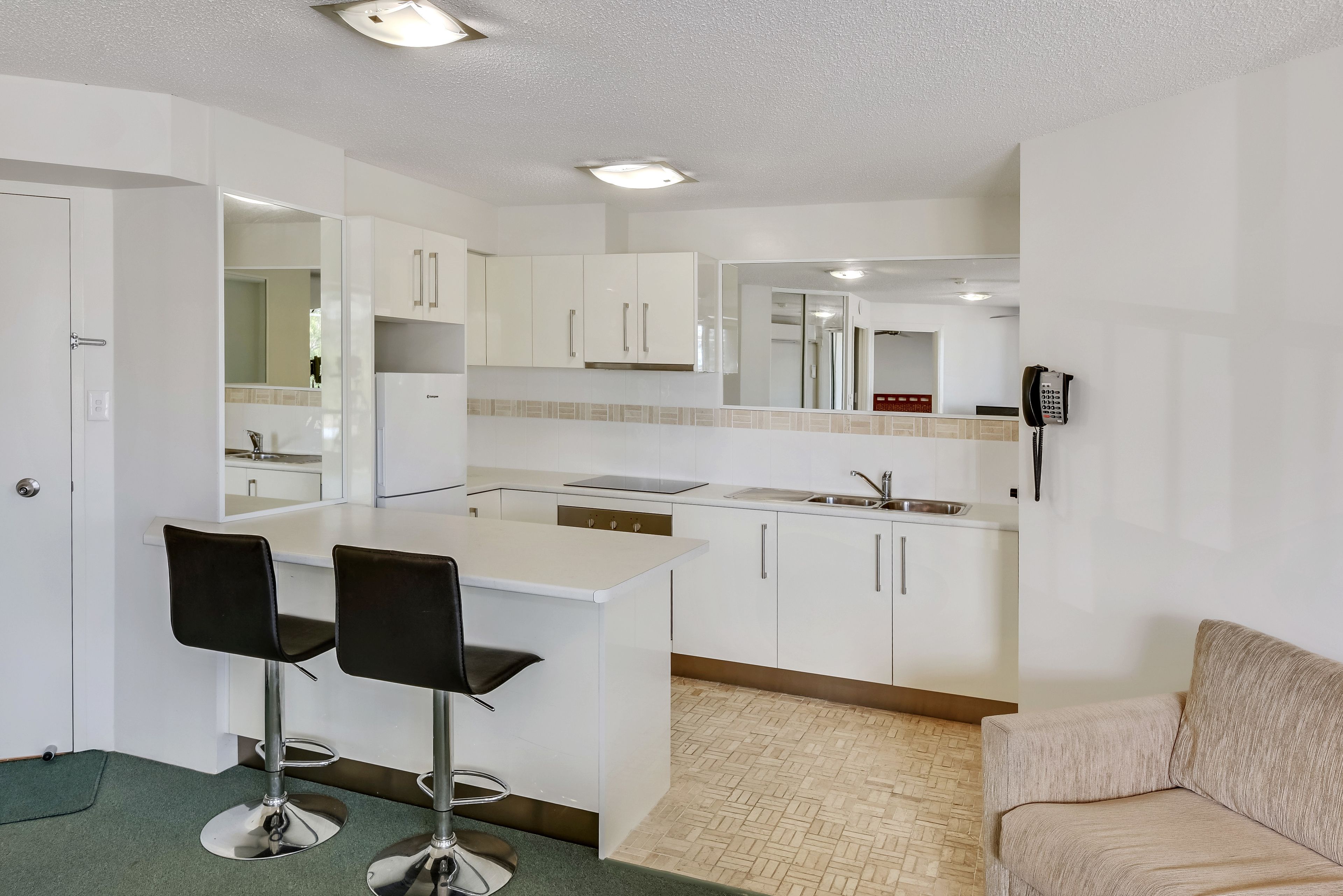 Una cocina moderna con armarios blancos, una isla central con dos taburetes de bar, un horno empotrado y un teléfono en la pared.
