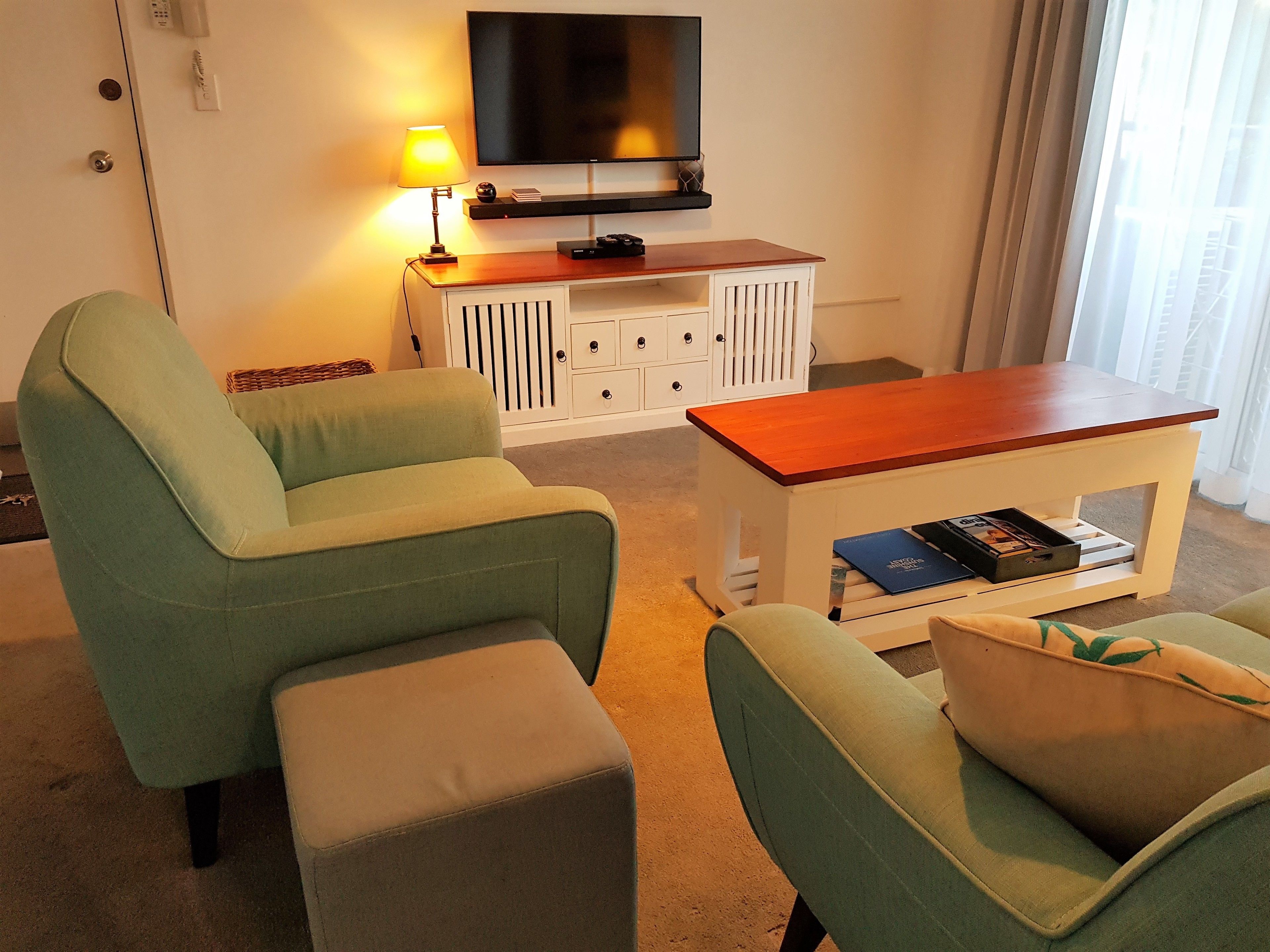 Una sala de estar con dos sillones verdes, una mesa de centro de madera, un mueble blanco con cajones, un televisor de pantalla plana montado en la pared y una lámpara sobre la mesa.